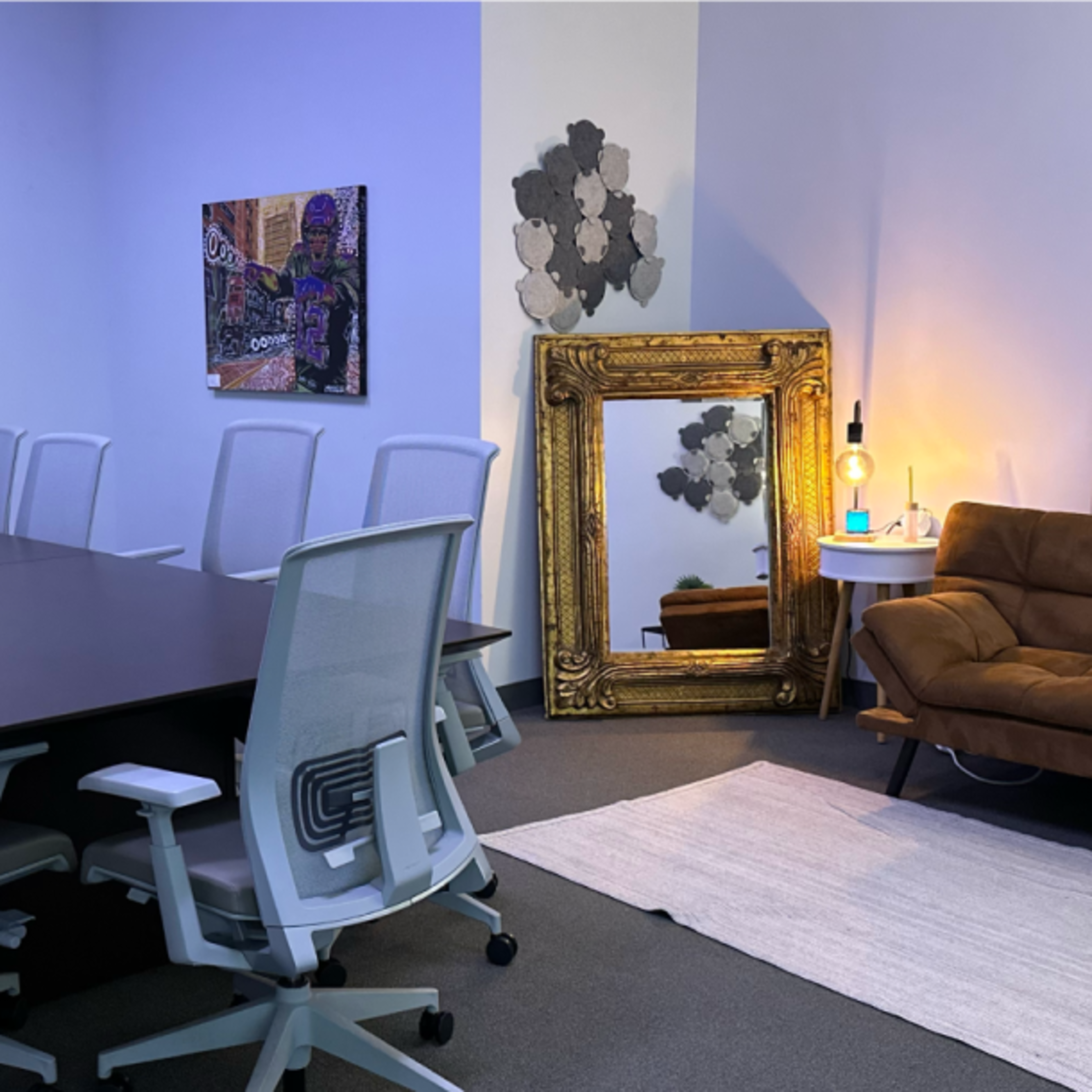 A modern conference room featuring a long table surrounded by chairs, a large screen on the wall, a decorative mirror, and a brown couch.