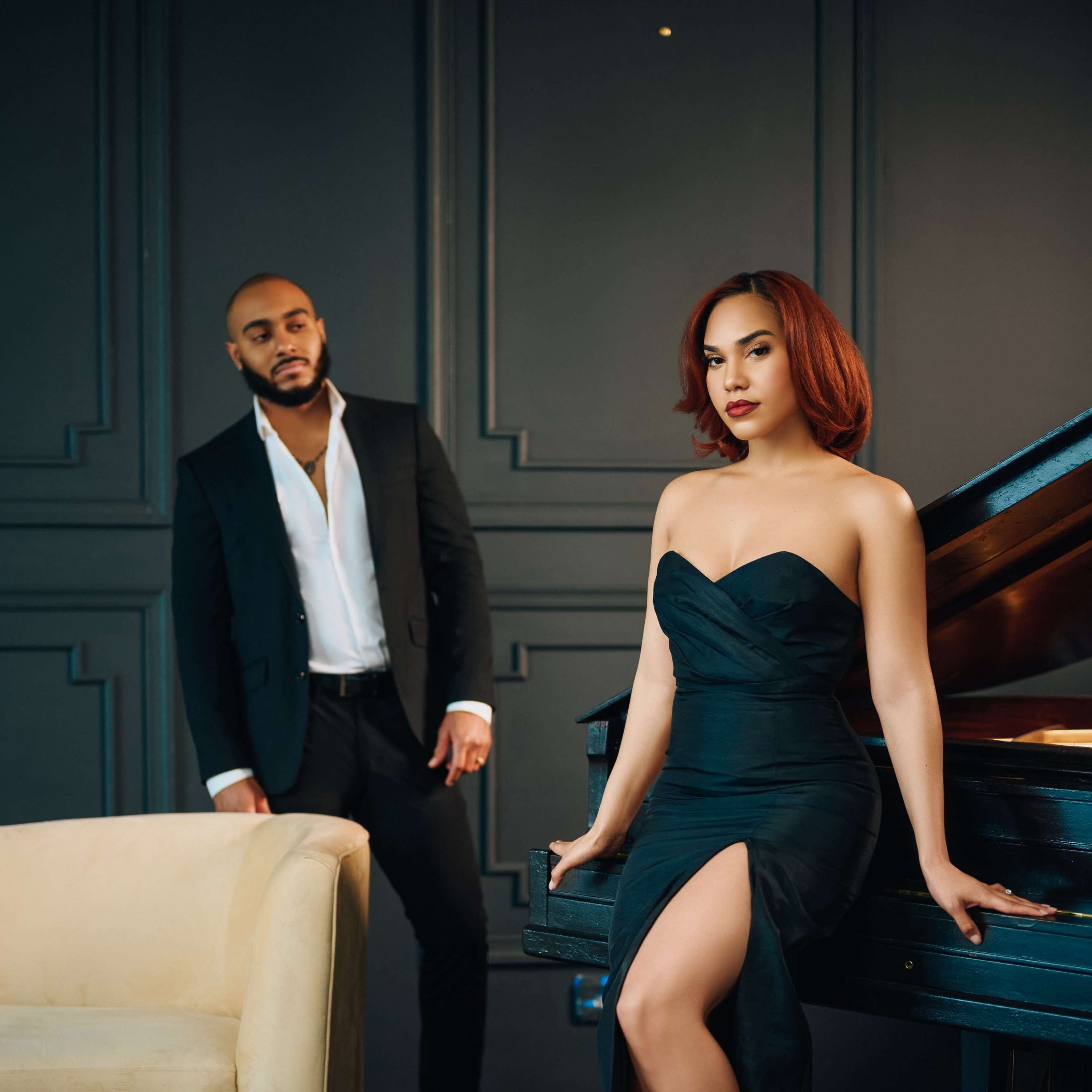 A woman in a black dress poses by a grand piano while a man in a suit stands nearby.