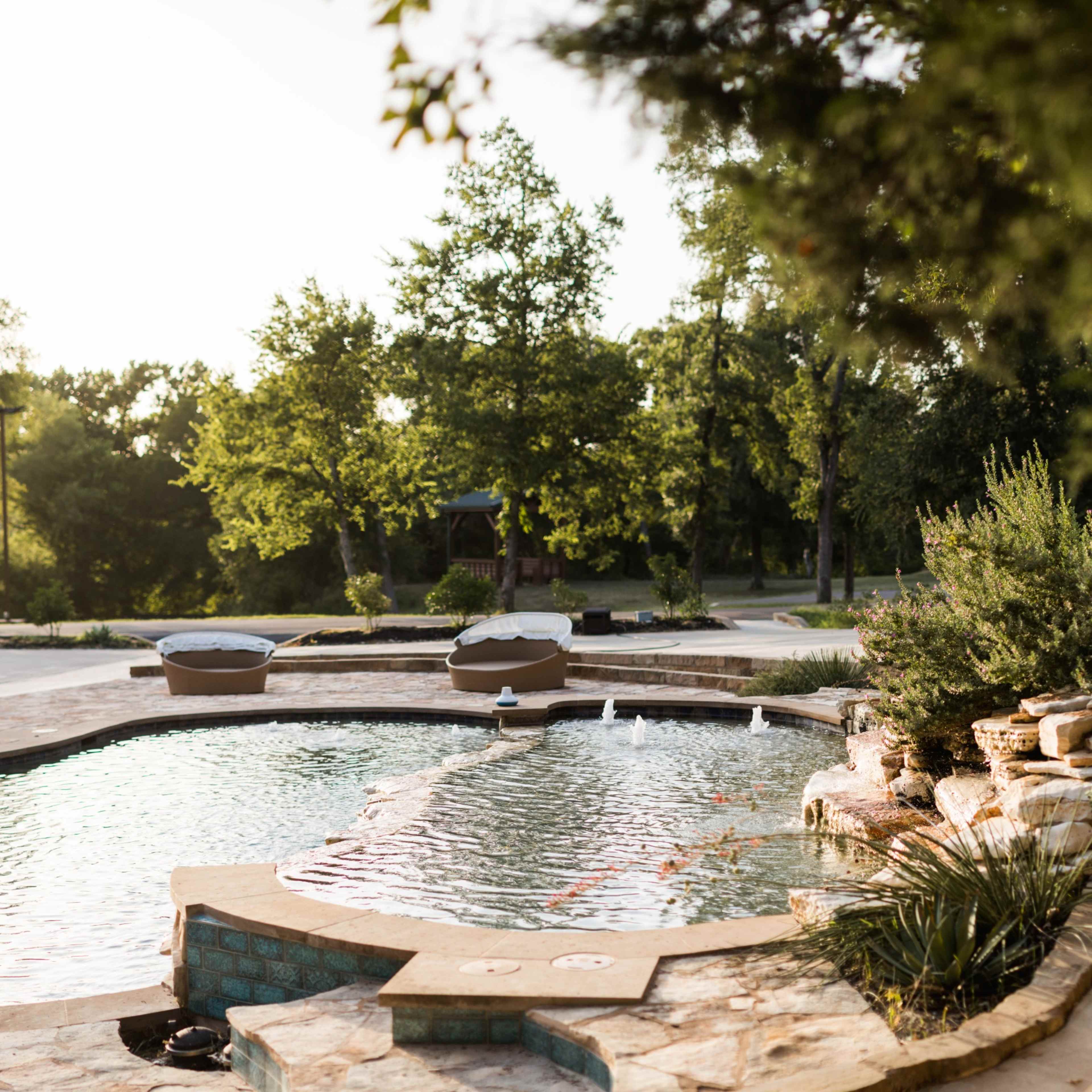 A curved swimming pool with stone edging is surrounded by lush greenery and sunlight filtering through the trees.