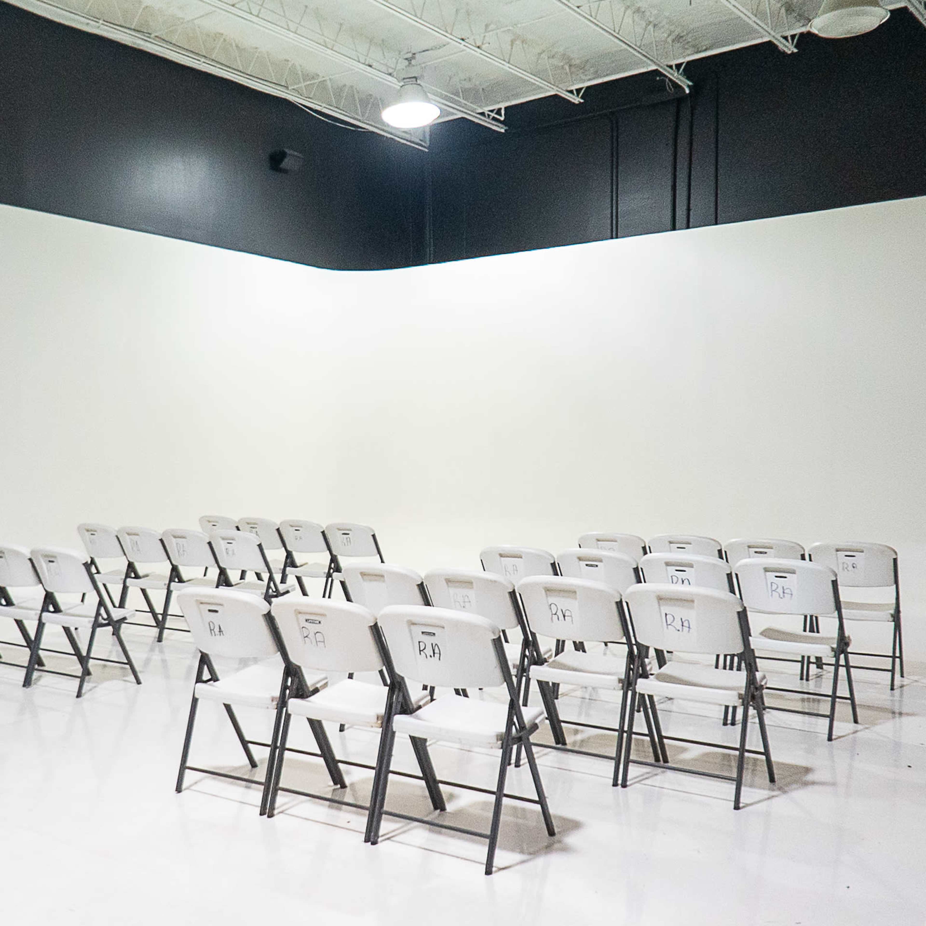 A row of white folding chairs is arranged facing a blank wall in a spacious, brightly lit room.