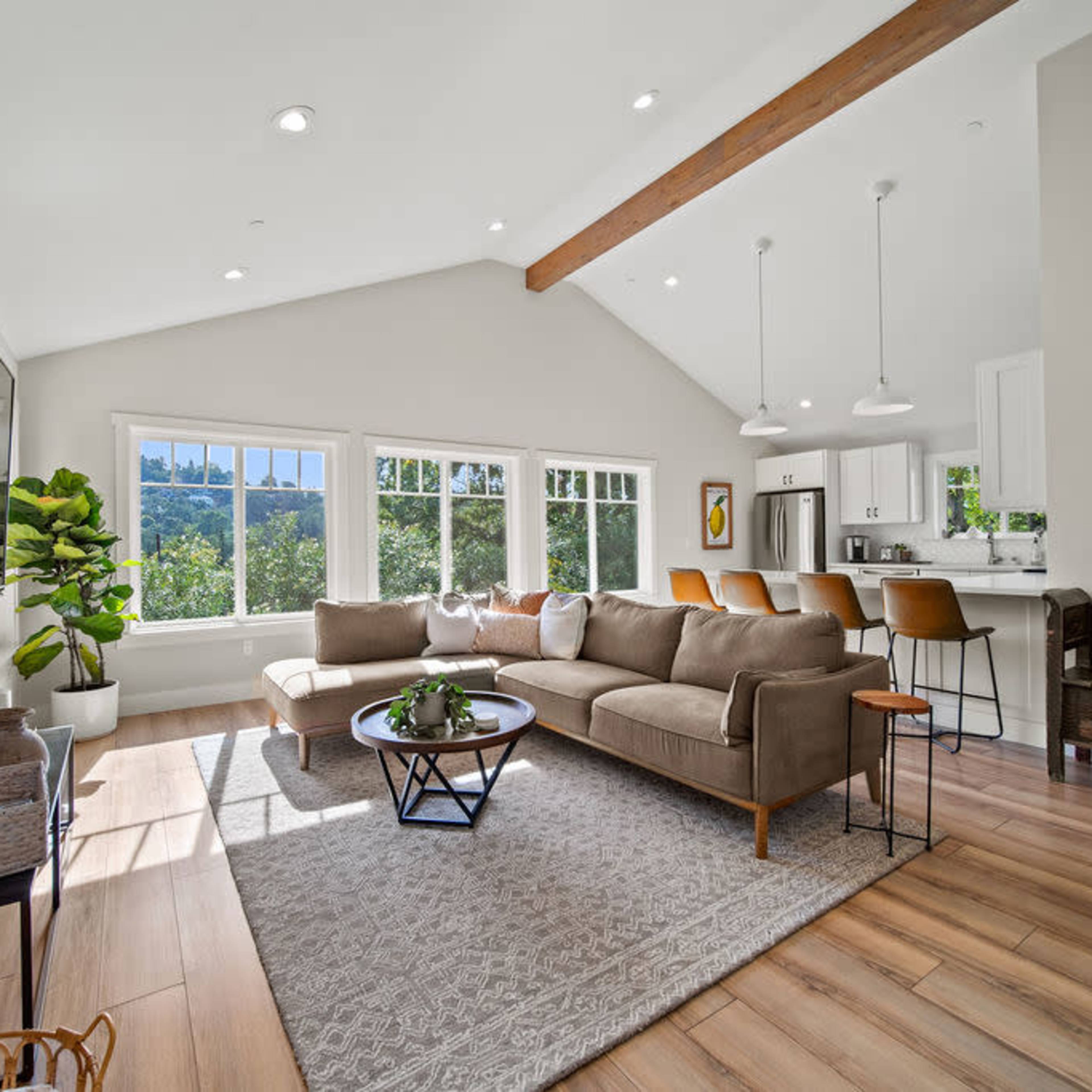 The living room features a beige sofa, a coffee table, and large windows that provide a view of the outdoors, with a kitchen visible in the background.