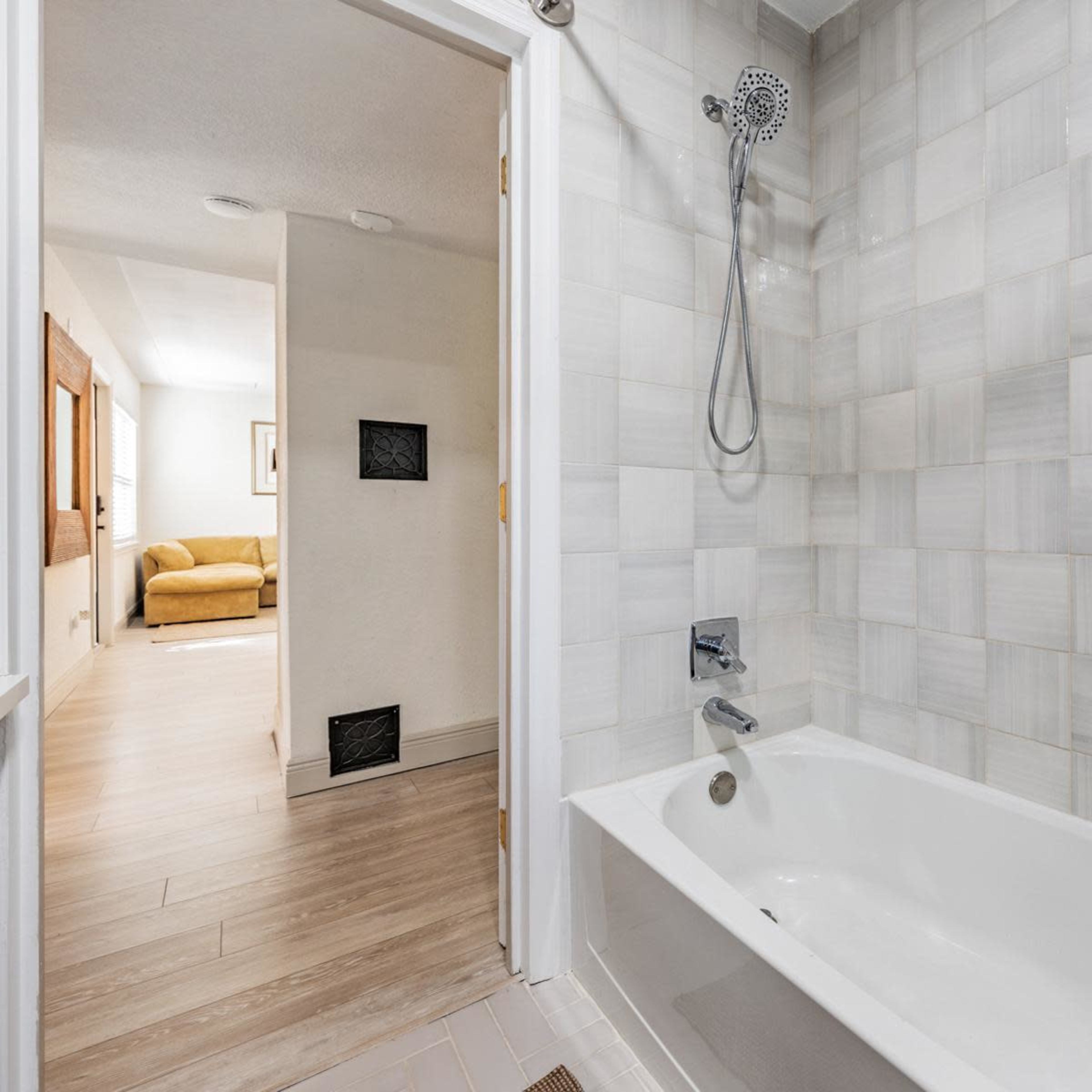 A bathroom featuring a white bathtub, a showerhead mounted on the wall, and gray tiled walls, with a doorway leading to a living space in the background.