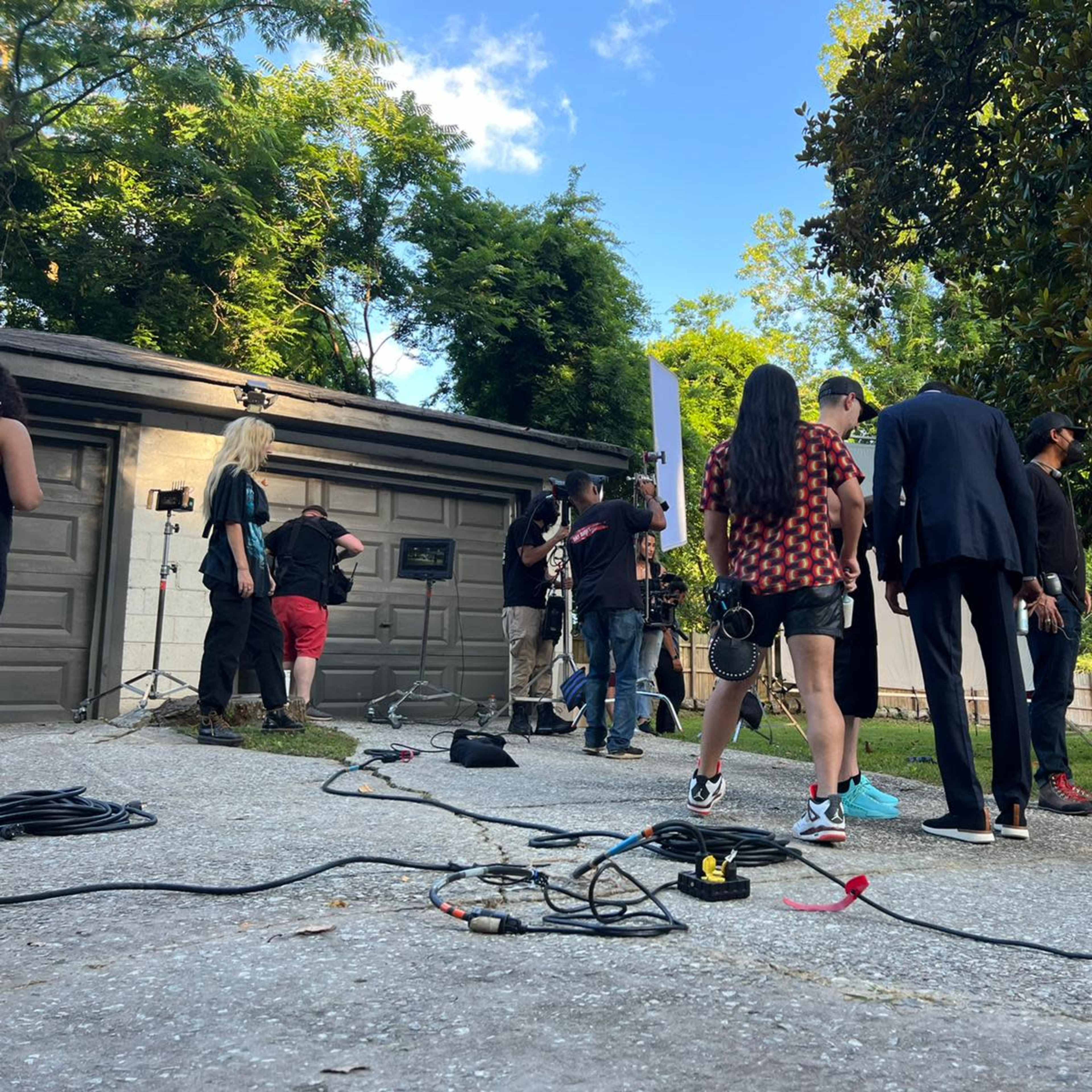 A group of people is gathered outside a garage set up for a filming production, with cameras and lights positioned around them.