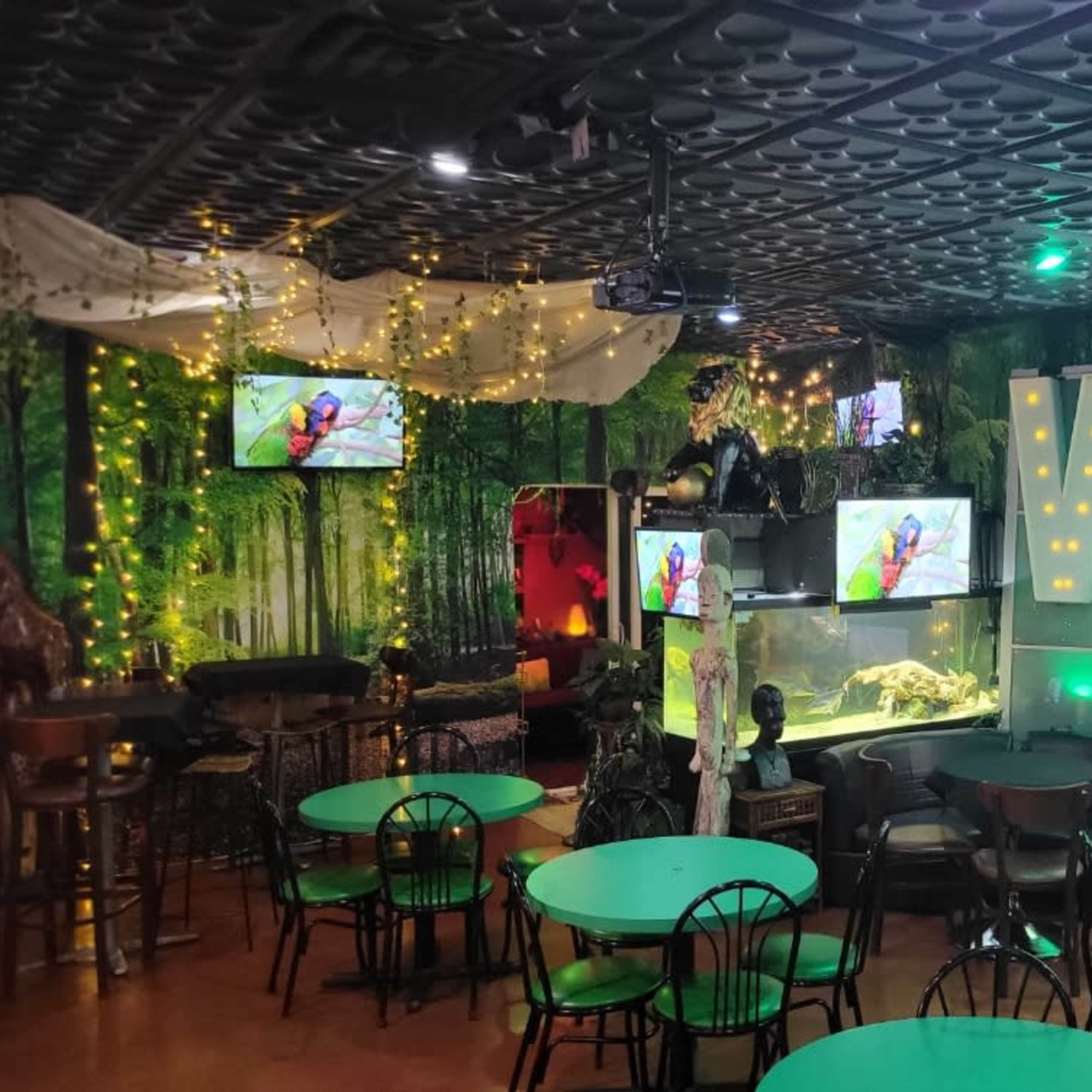 A themed café or restaurant decorated with lush greenery, multiple screens displaying nature scenes, aquariums, and a seating area with tables and chairs.