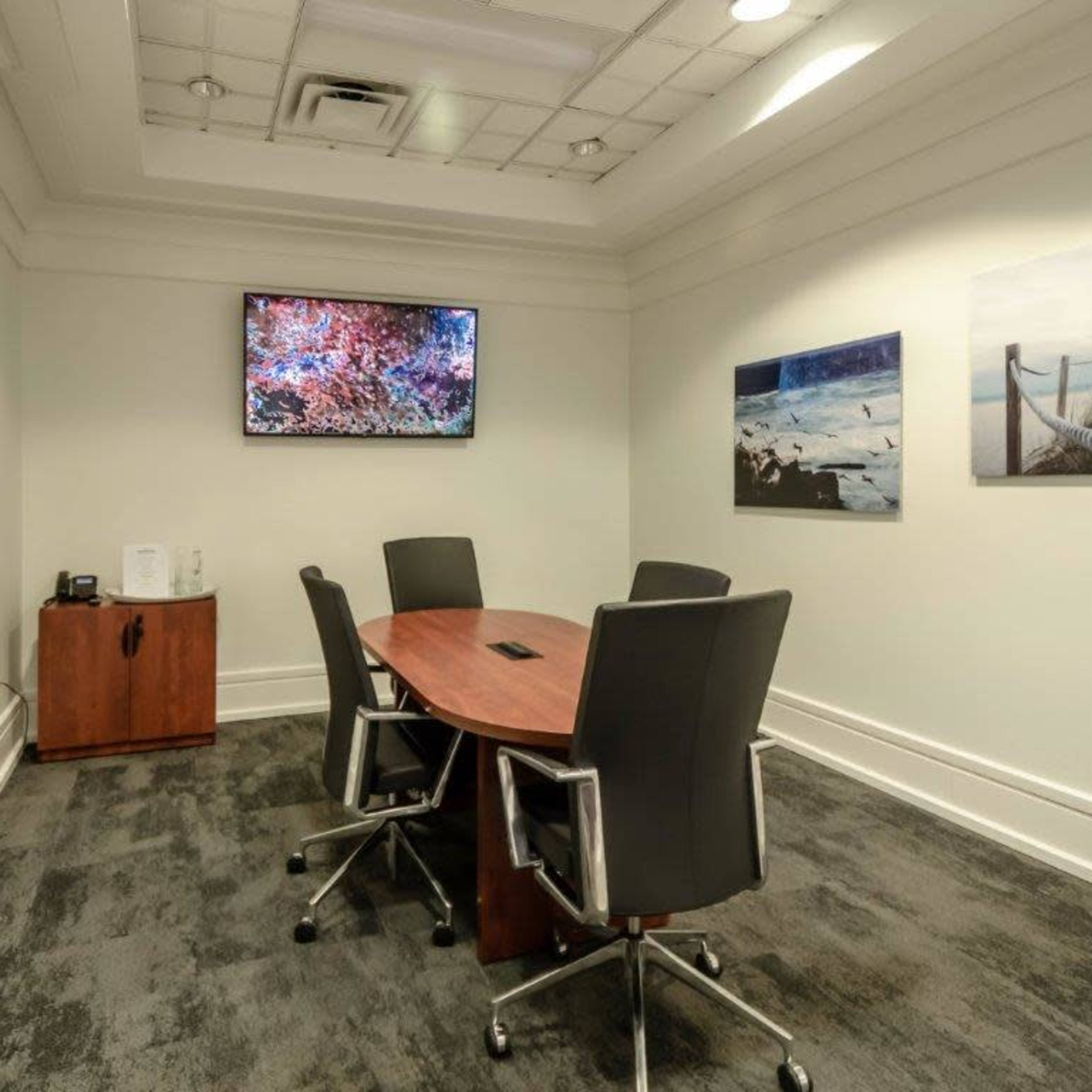 A small conference room features a round wooden table with four ergonomic chairs, a wall-mounted television, and two framed landscape photos.