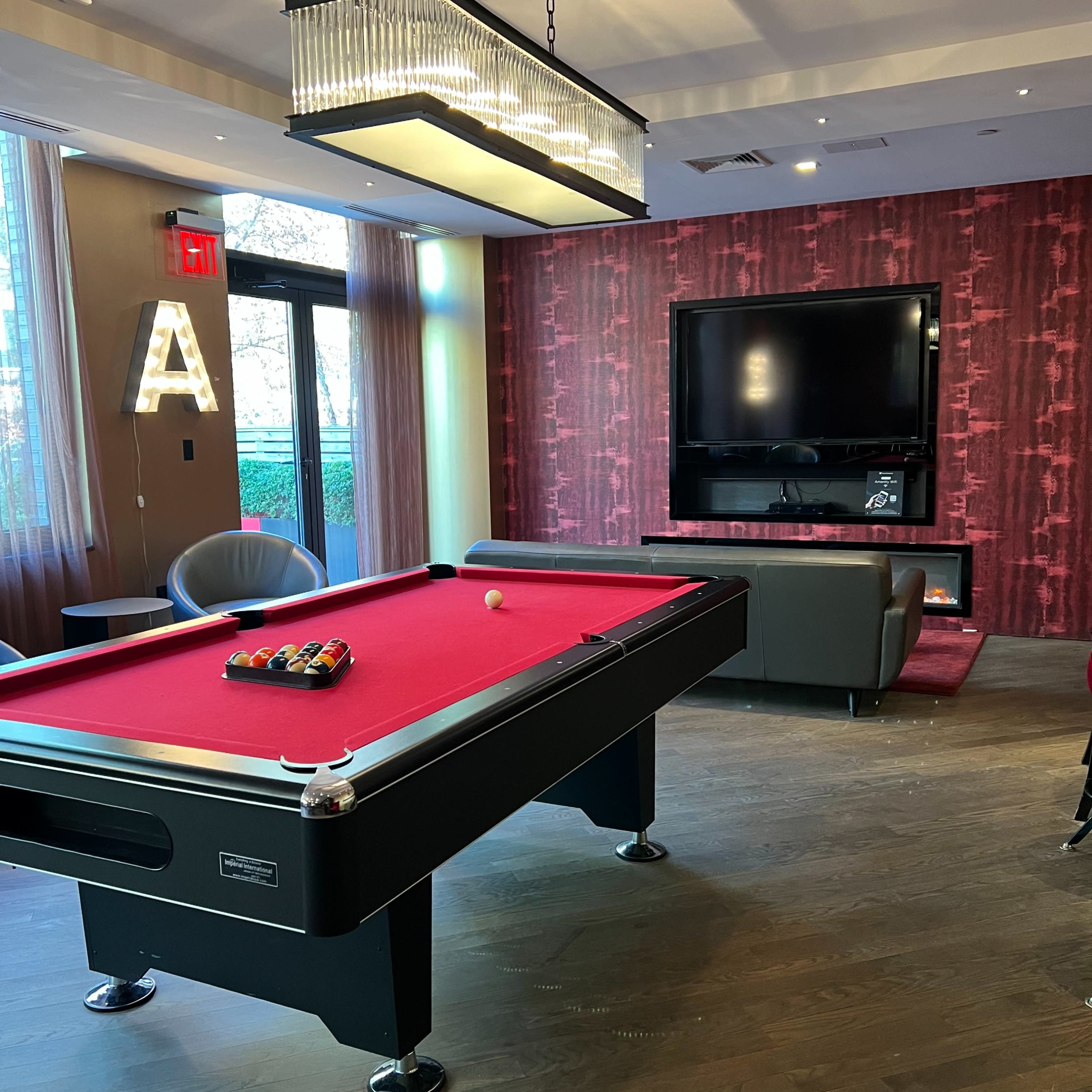 A modern game room featuring a pool table with a red felt surface, a large flat-screen TV on a decorative wall, and a stylish lamp hanging overhead.