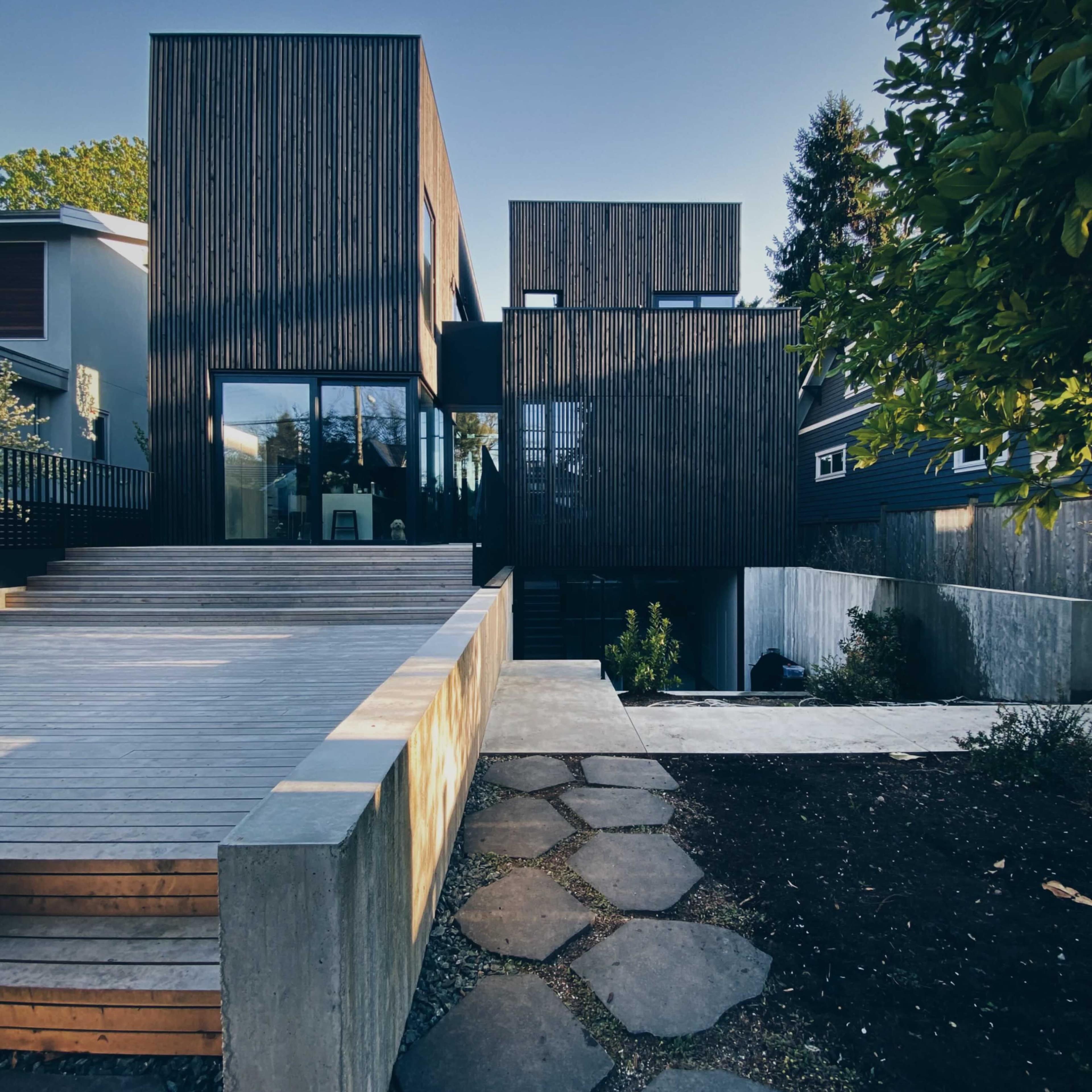 A modern wooden-clad house with large windows, surrounded by a landscaped yard featuring stone pathways and a deck.