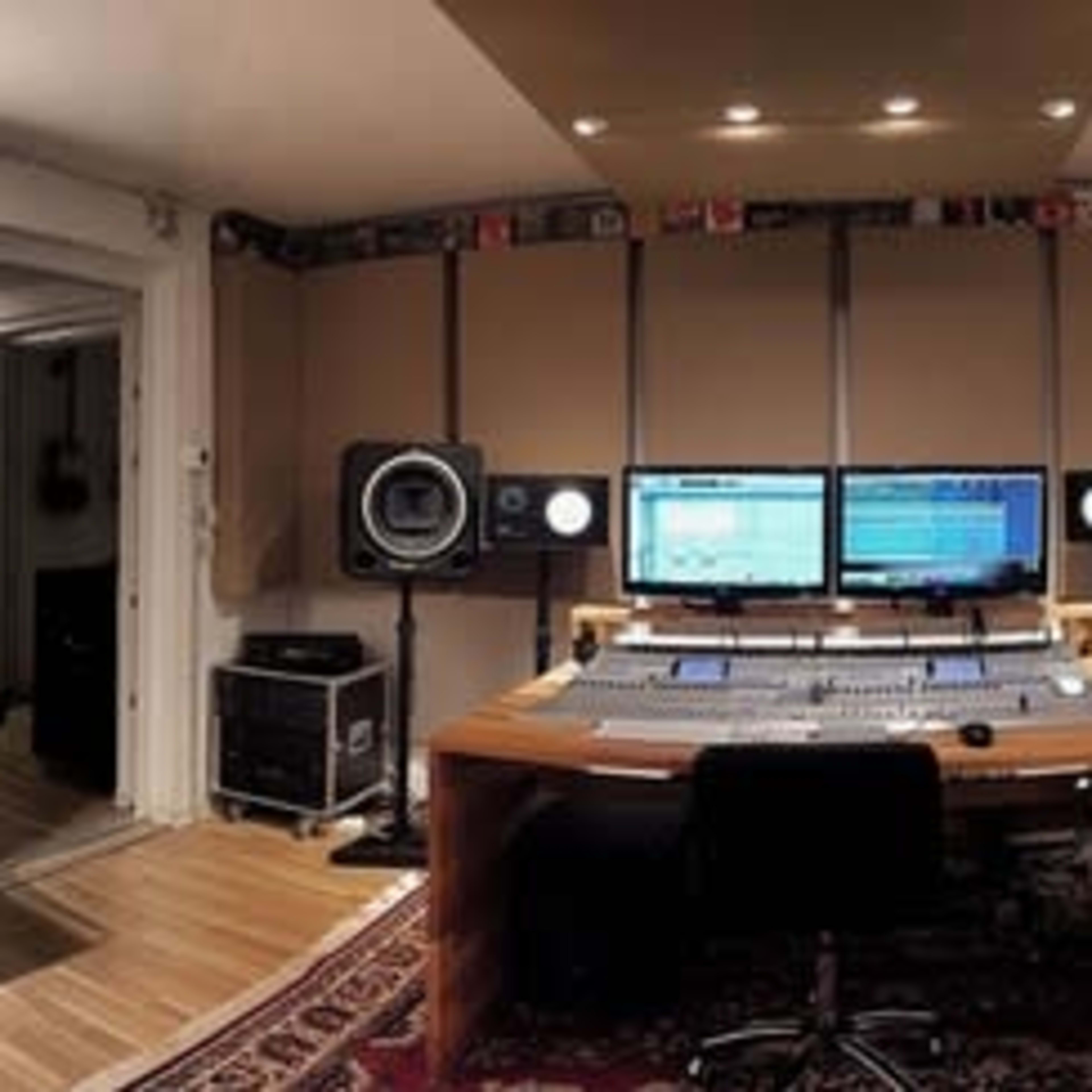 A music studio featuring a large mixing desk, two computer monitors, speakers, a keyboard, and various audio equipment in a well-furnished setting.