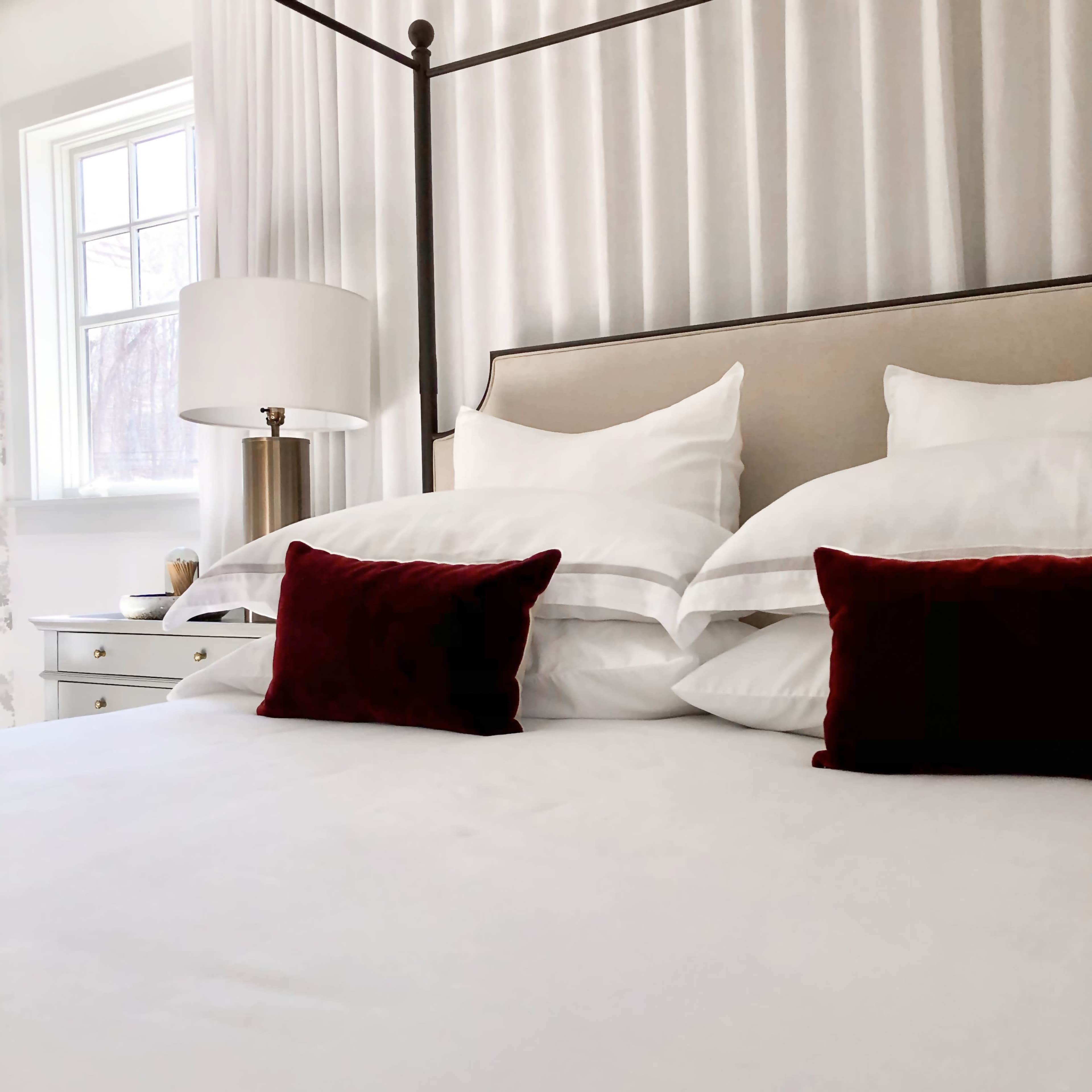 A neatly made bed with white linens and two red accent pillows is featured in a softly lit bedroom.