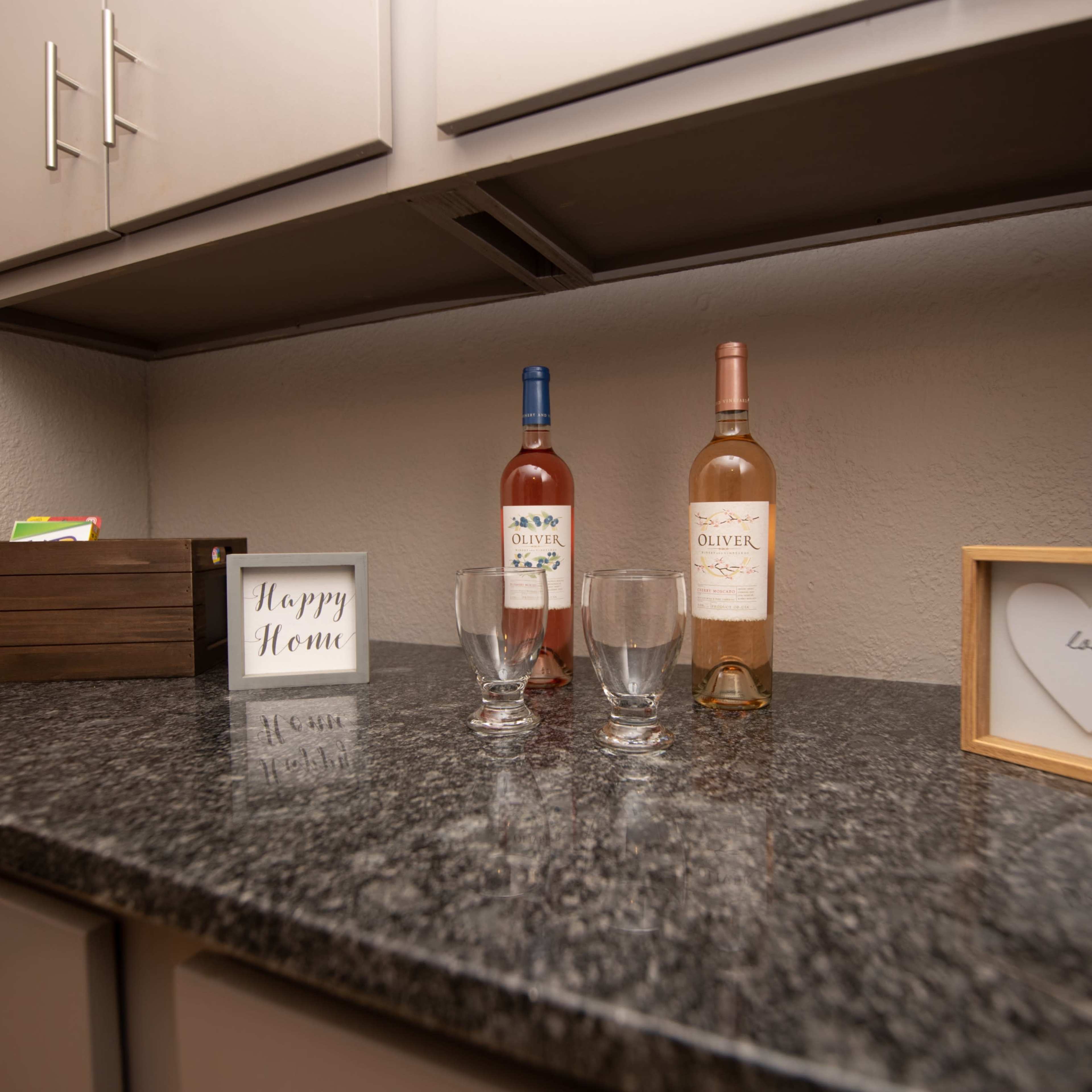 A wooden countertop displays two bottles of wine, glasses, a box, and framed decorative items.