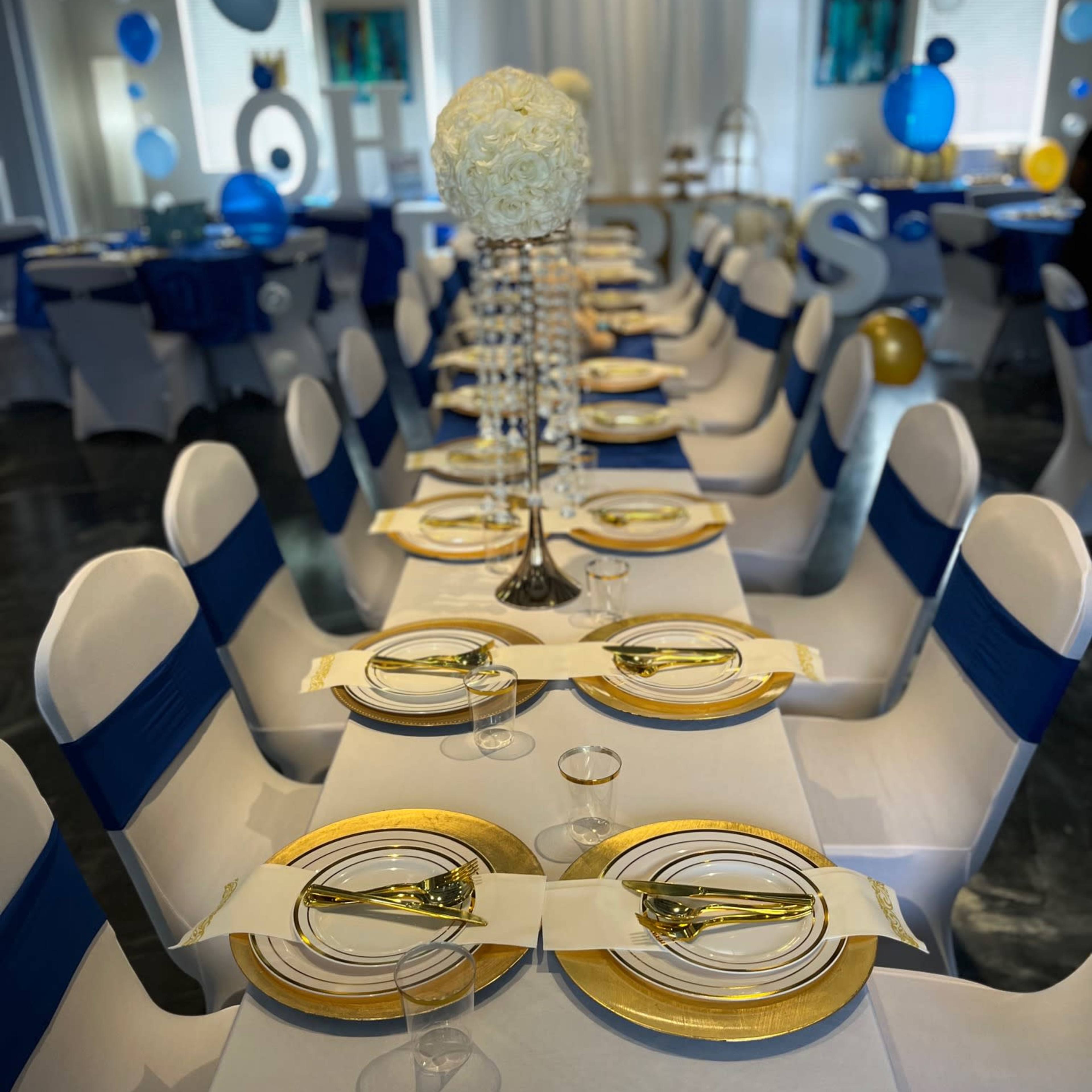 A long banquet table is elegantly set with gold-rimmed plates, sparkling glassware, and white floral centerpieces, surrounded by chairs covered in blue and white fabric.