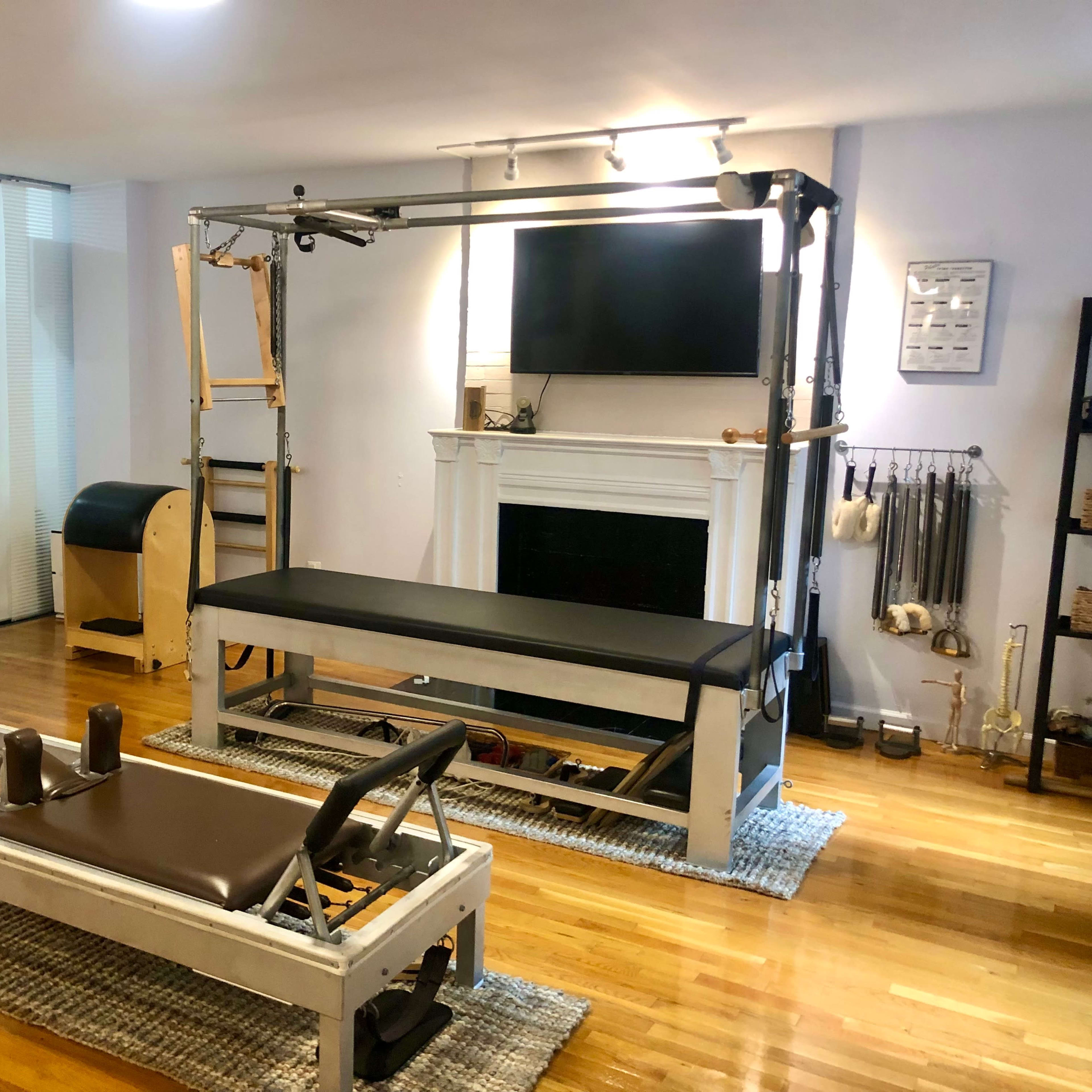 A neatly organized indoor studio featuring a reformer Pilates machine, an elevated Pilates platform, and various fitness equipment along with a television mounted on the wall.