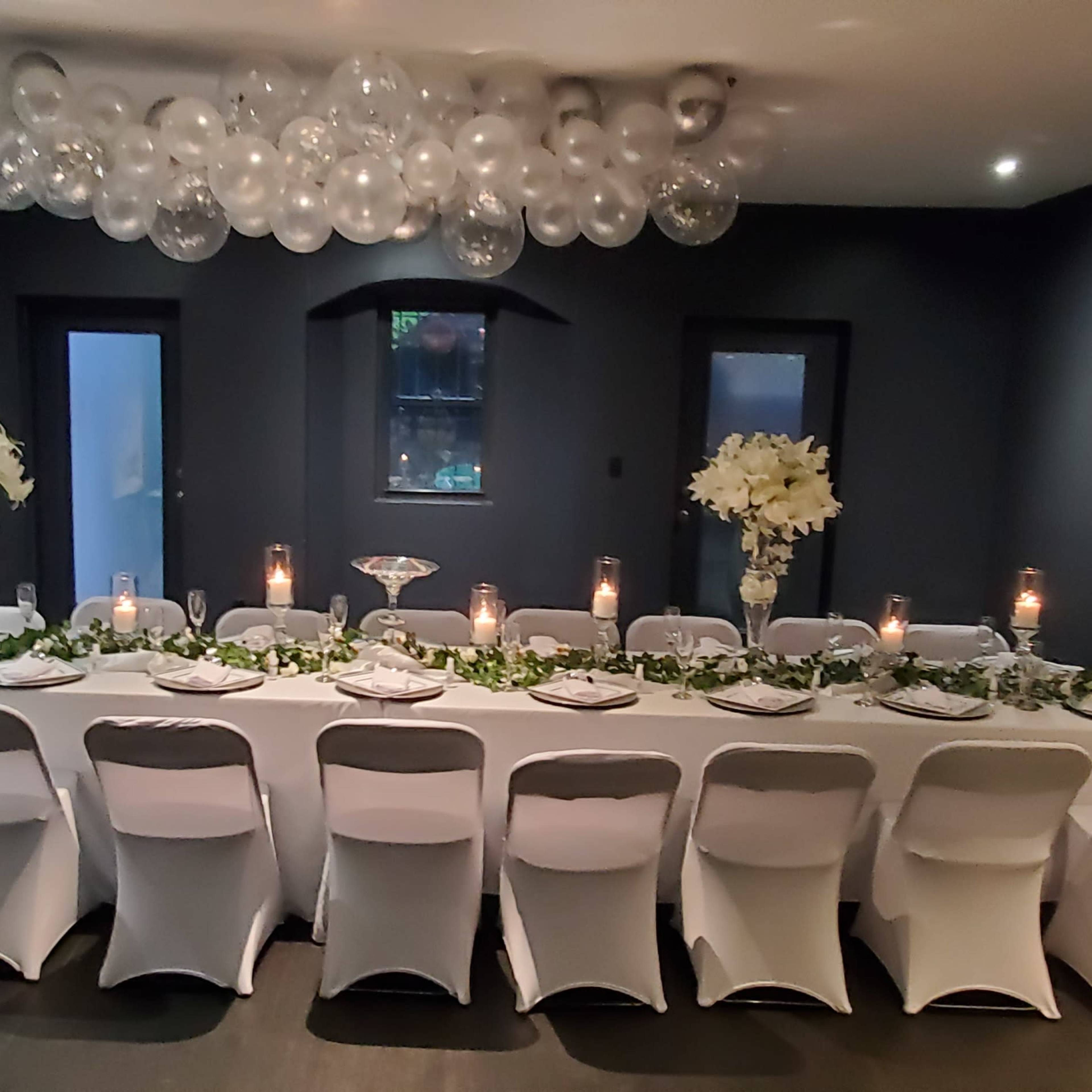 A long, elegantly set dining table with white chairs is arranged in a dark room, decorated with flowers, candles, and overhead glass orbs.
