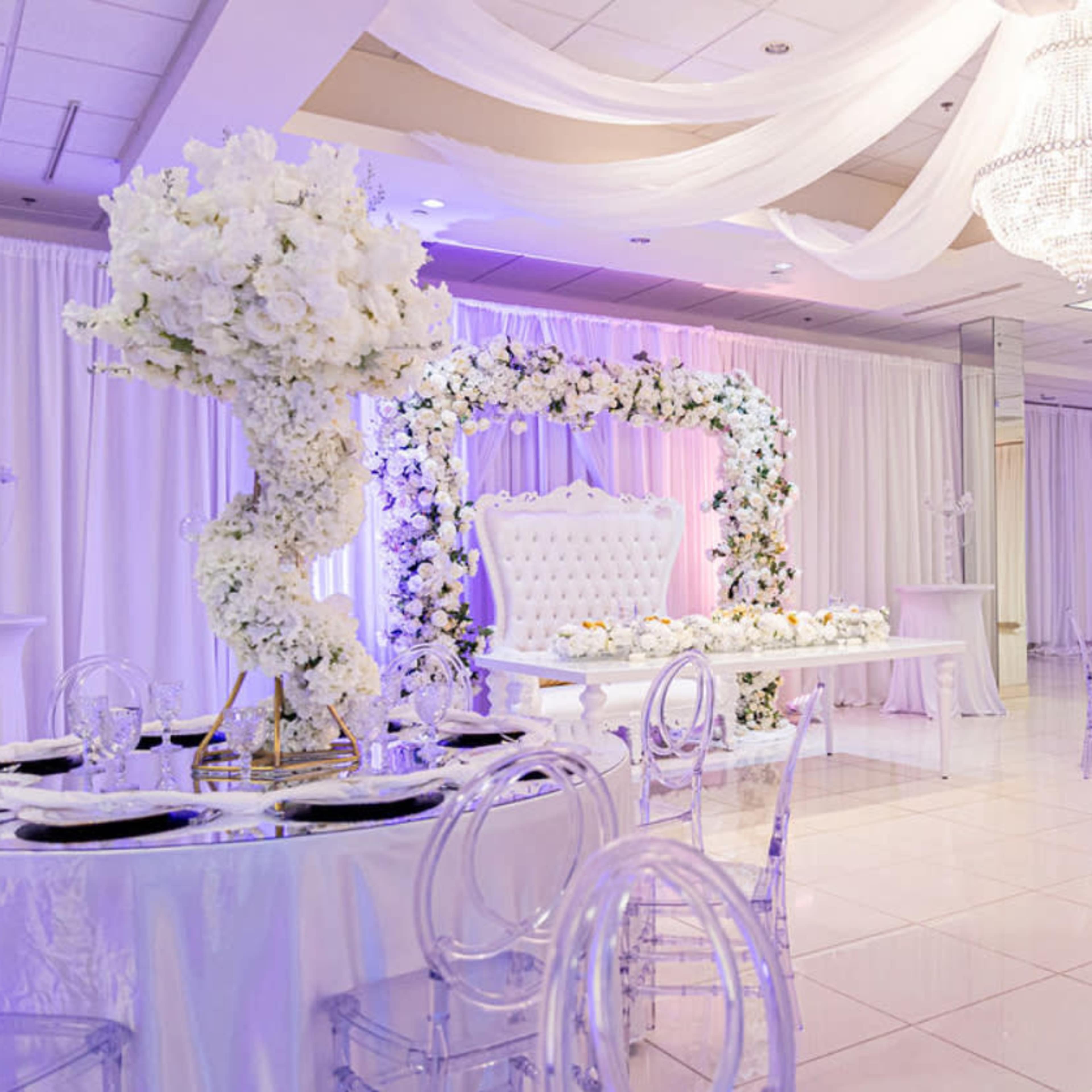 A beautifully decorated event space with white drapery, elegant floral arrangements, and crystal chandeliers.