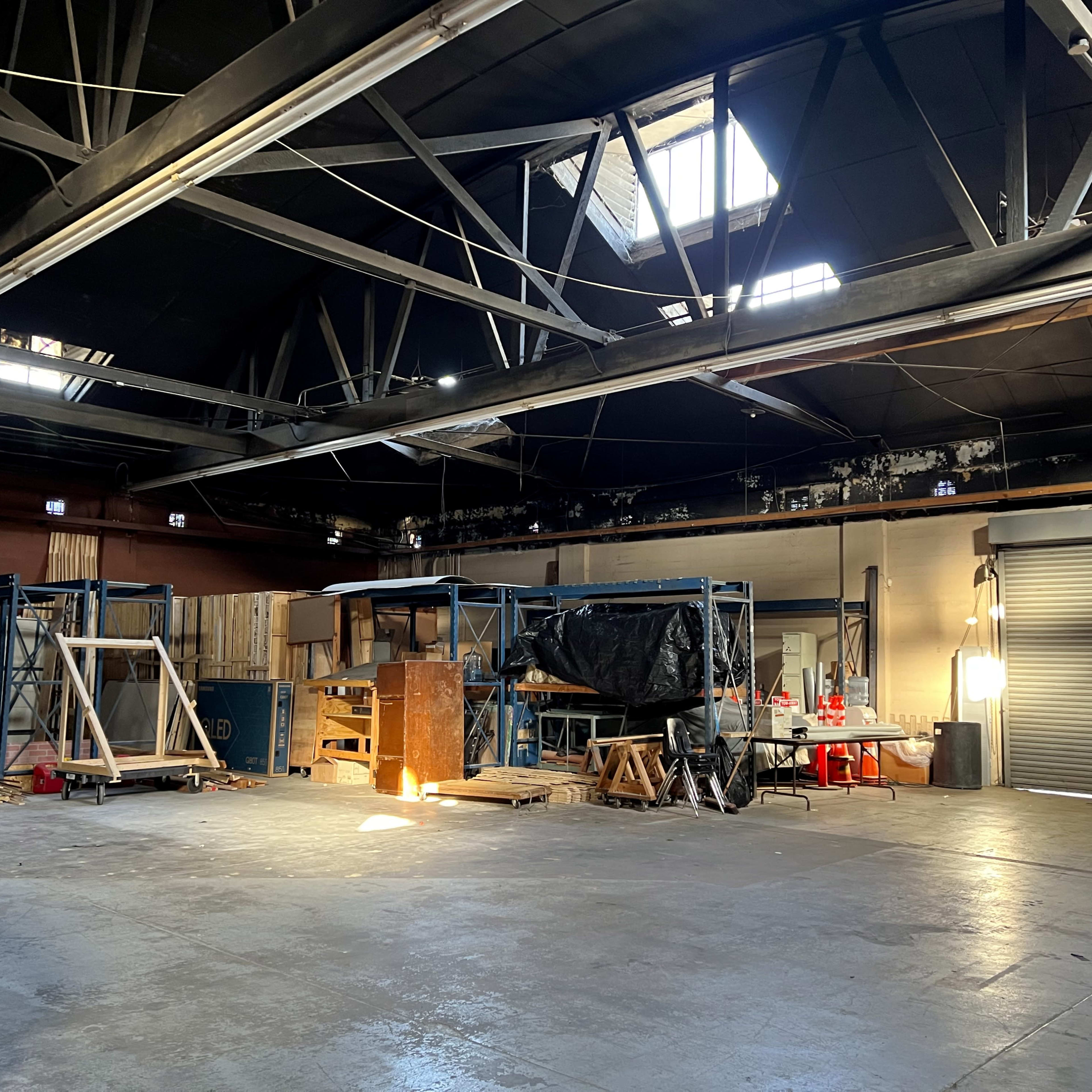 A large industrial space with storage shelves, wooden materials, and a red car parked nearby under natural light from a window.