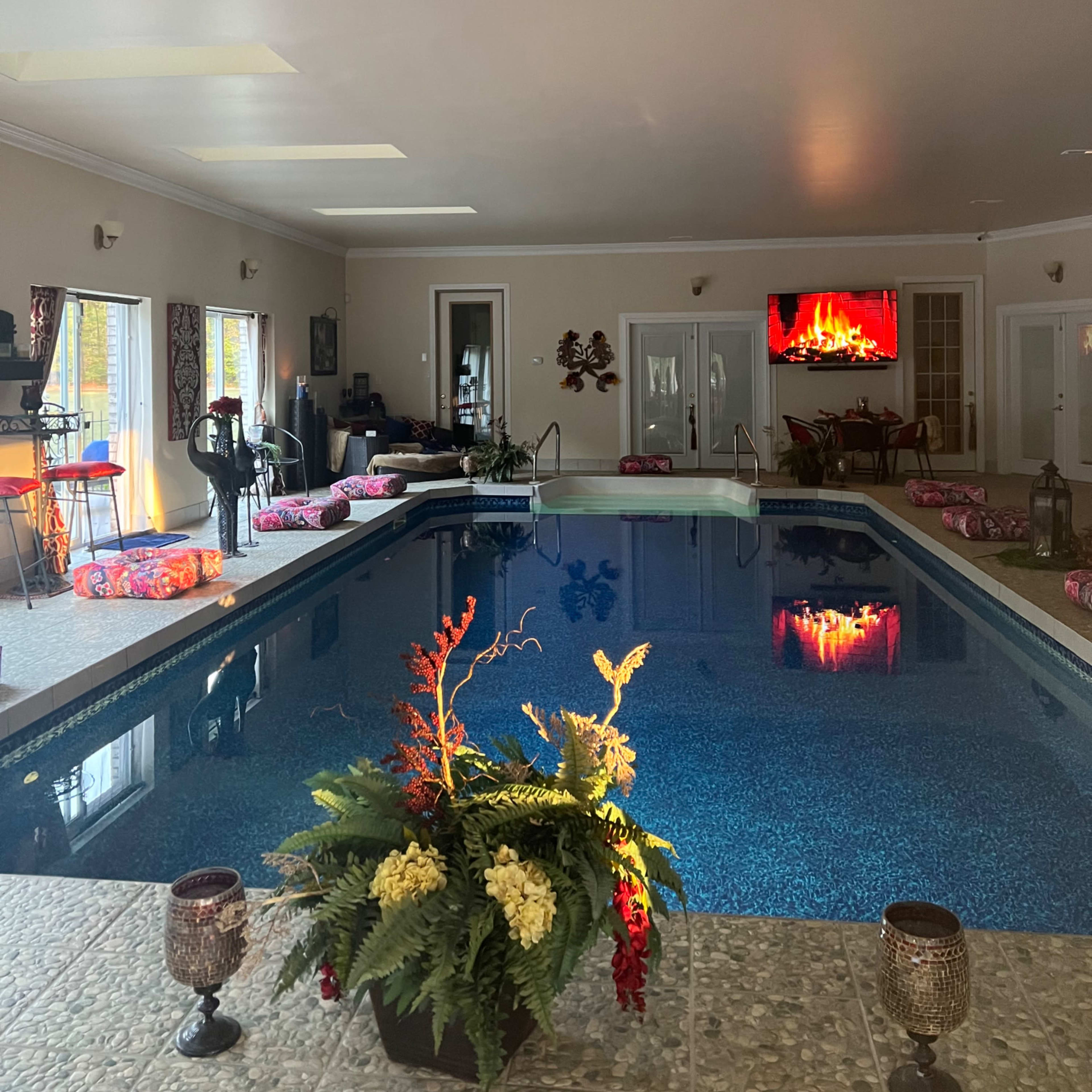 An indoor pool area with a large swimming pool, colorful seating arrangements, and a television displaying a fireplace.