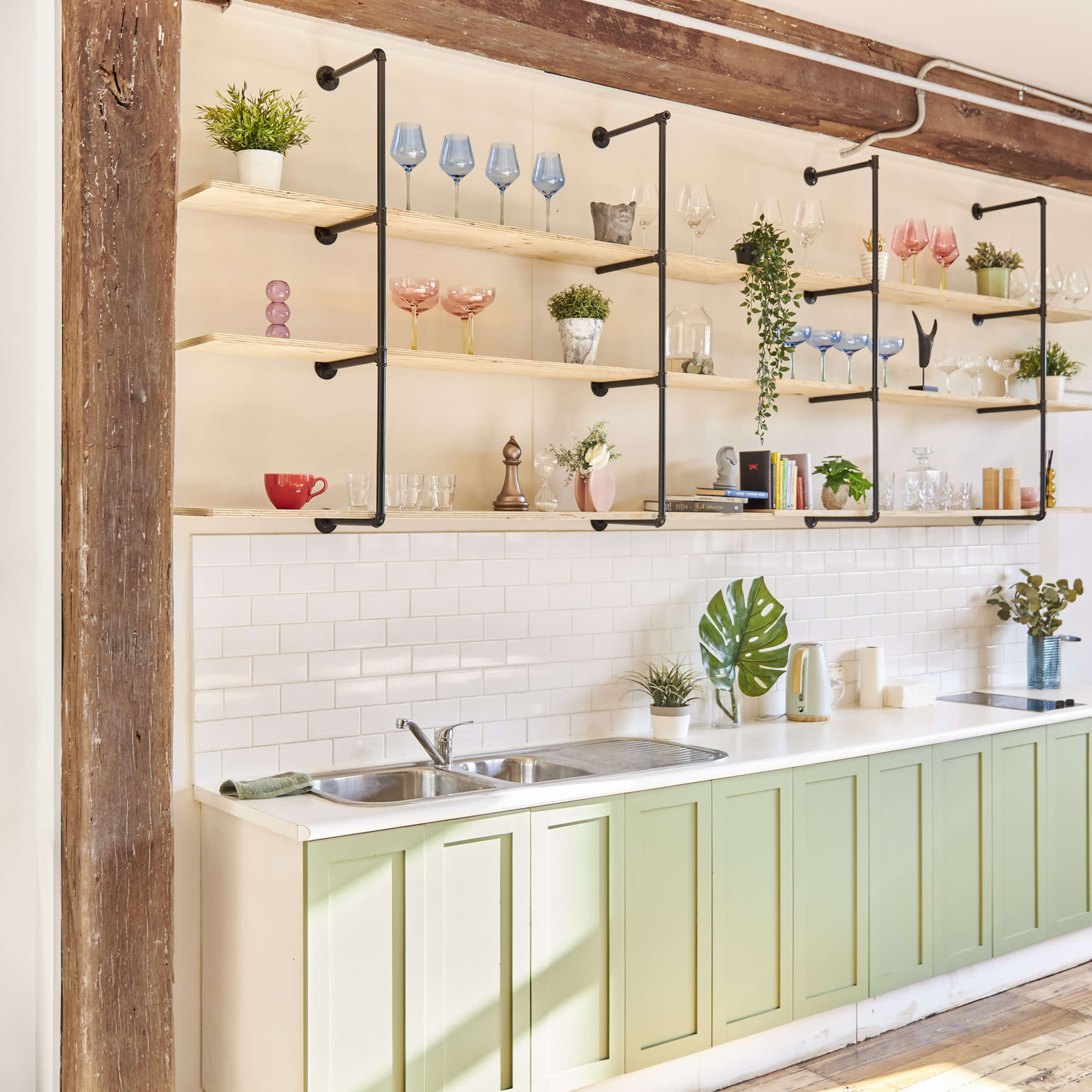A kitchen with a green cabinetry base, a white tiled backsplash, and open shelving displaying various glassware and potted plants.