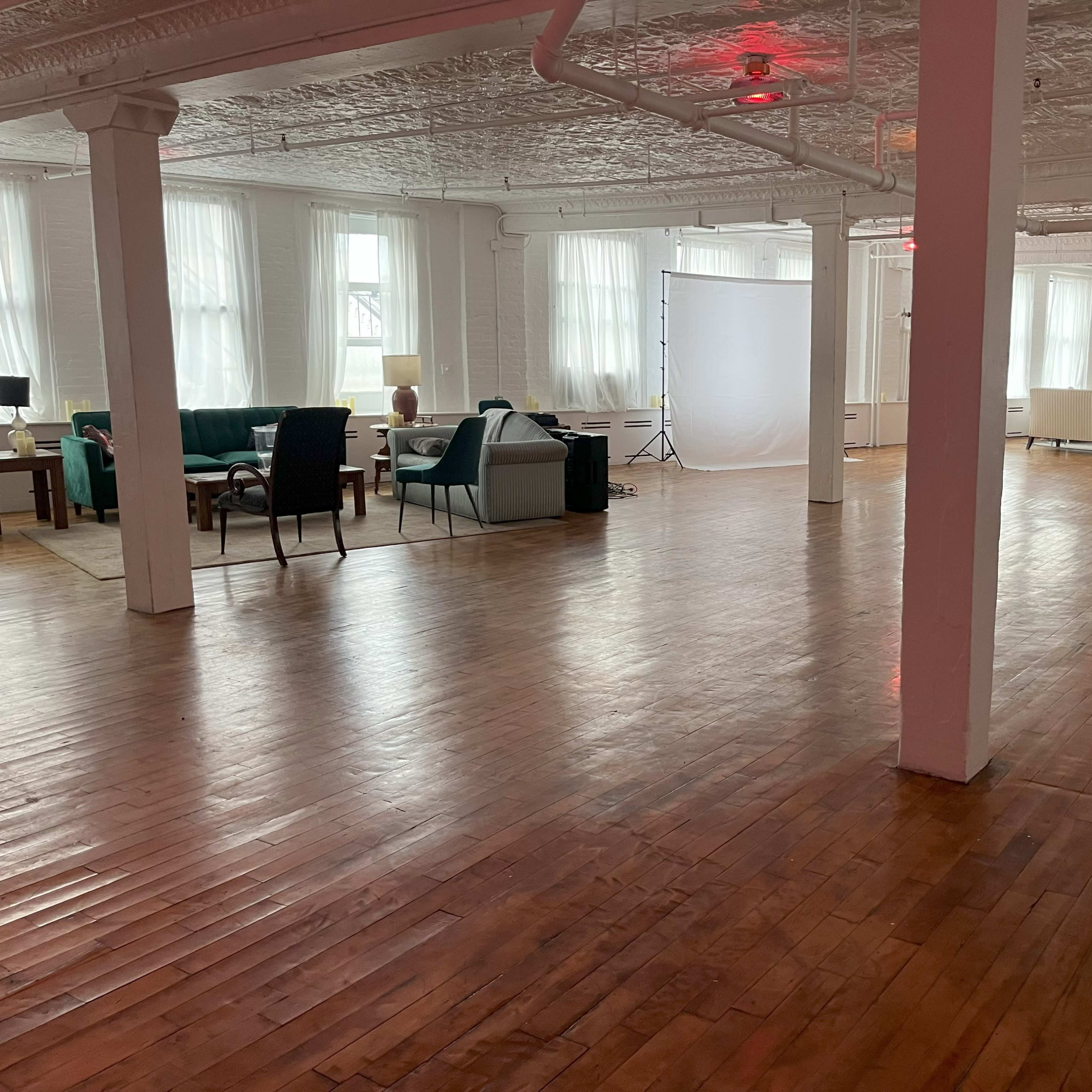 A spacious room with wooden floors, featuring various seating arrangements and bright natural light filtered through large windows.
