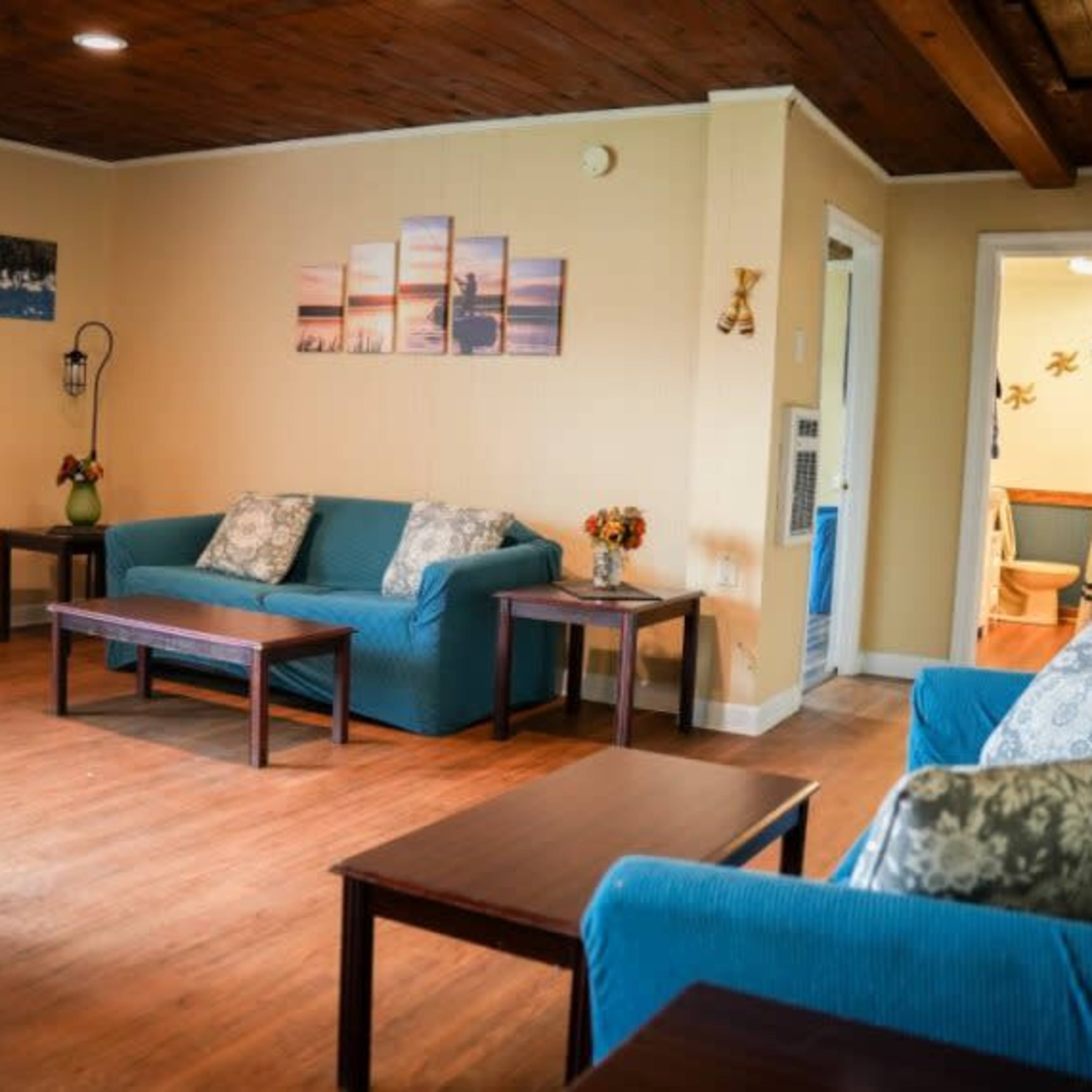 A living room with two blue sofas, wooden tables, and a doorway leading to a bathroom in the background.