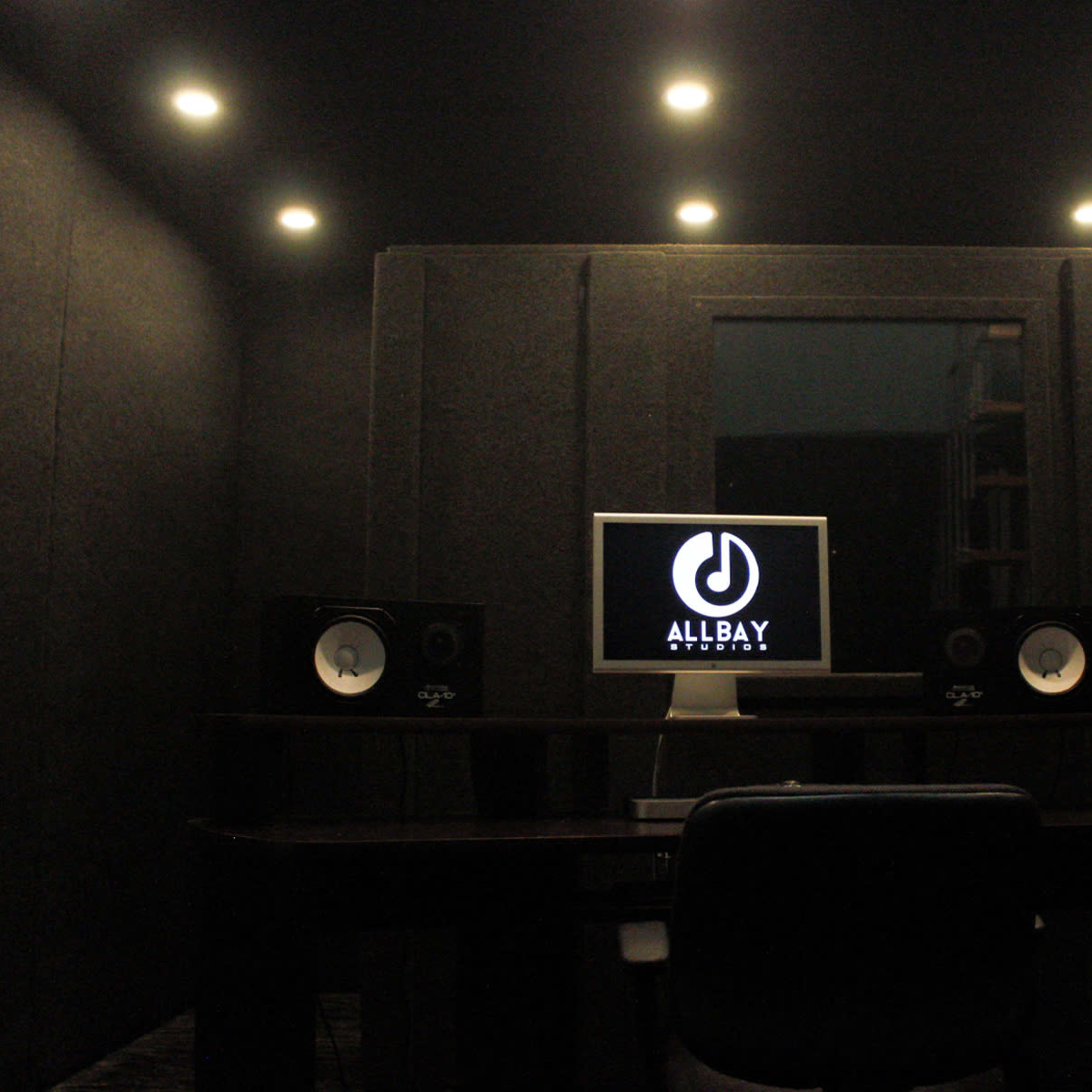 A soundproof recording studio features a computer monitor and speakers on a desk, with dark walls and ceiling lights.