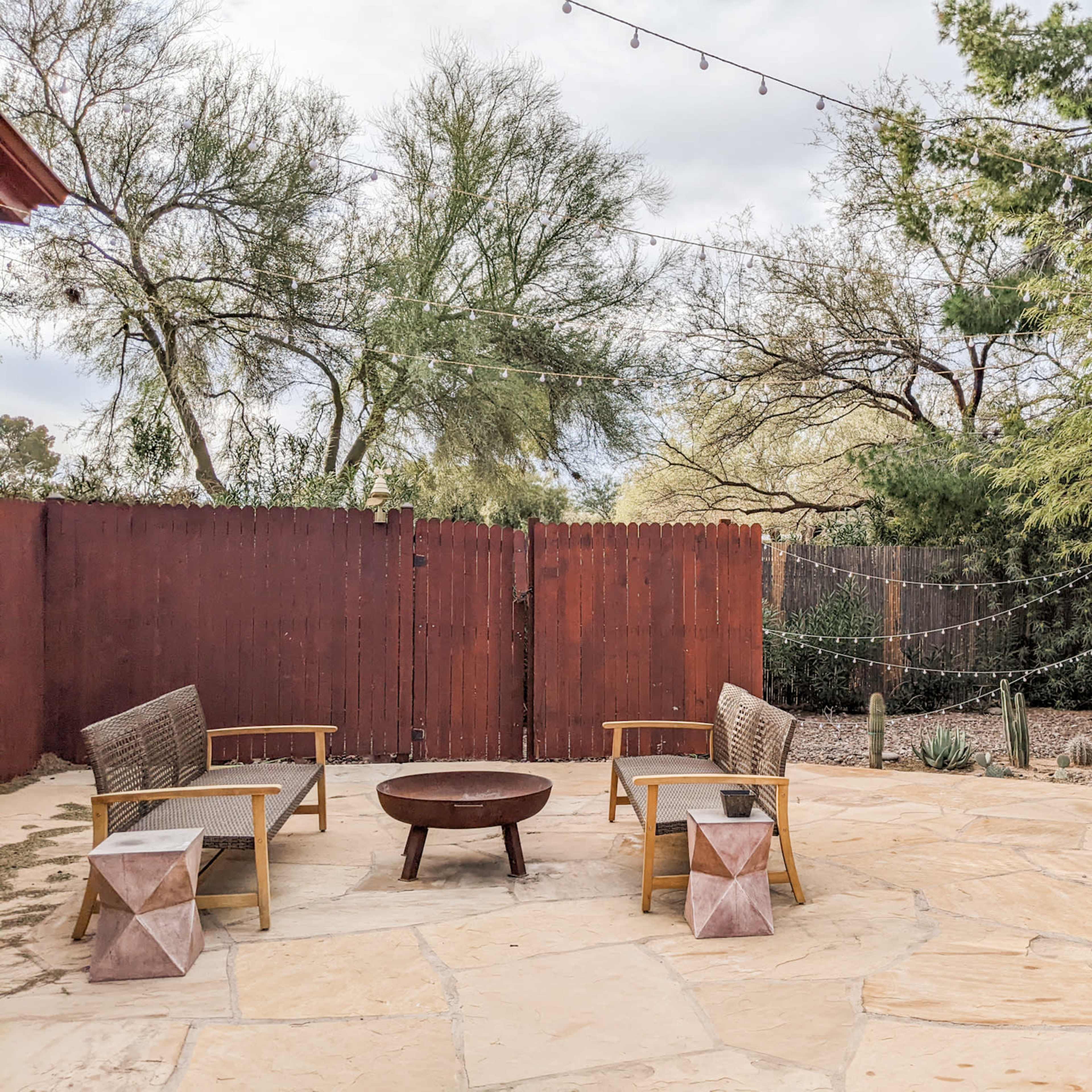 A stone patio features two chairs and a circular fire pit surrounded by trees and string lights.