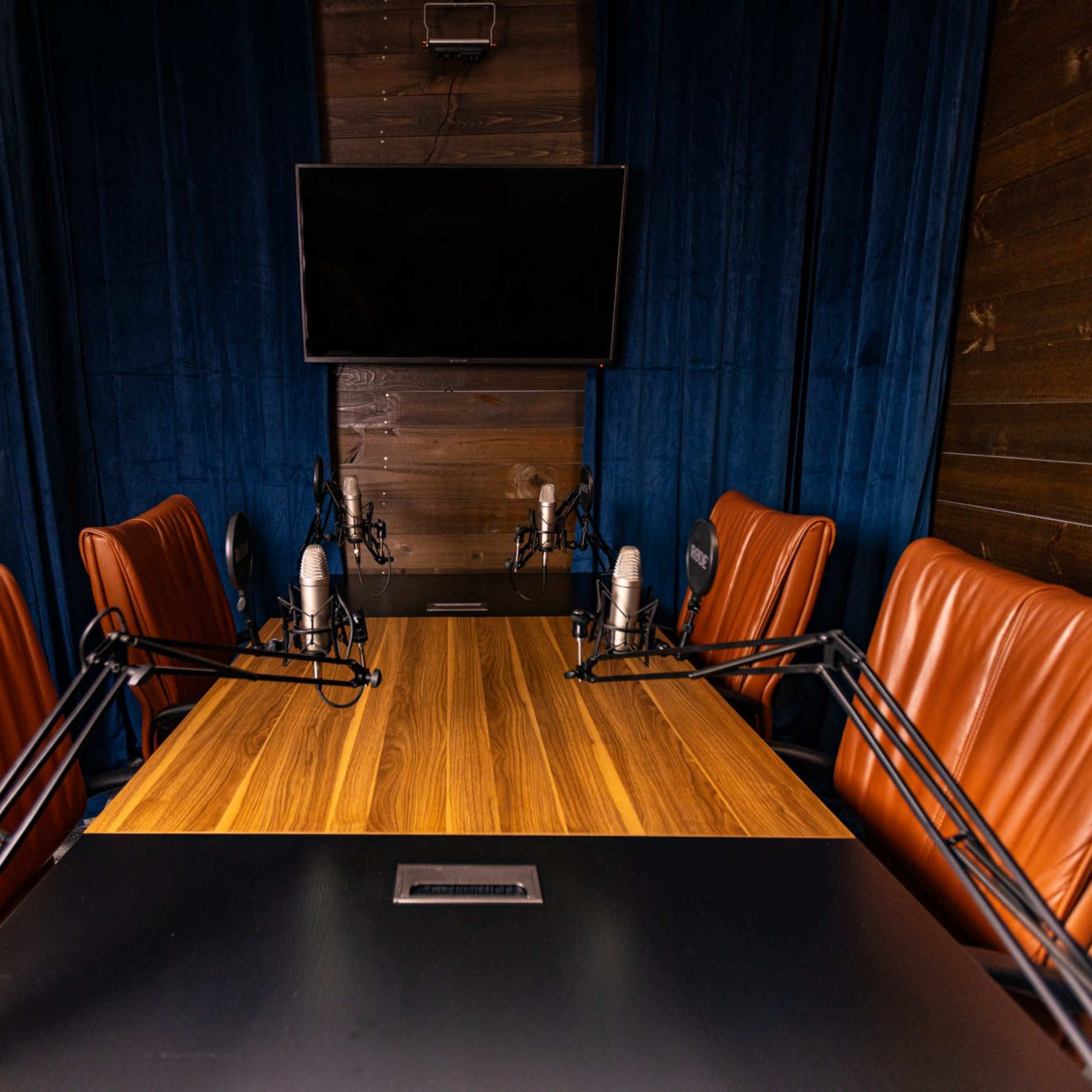 A conference room features a wooden table surrounded by four brown leather chairs and equipped with microphones, under a wall-mounted television.