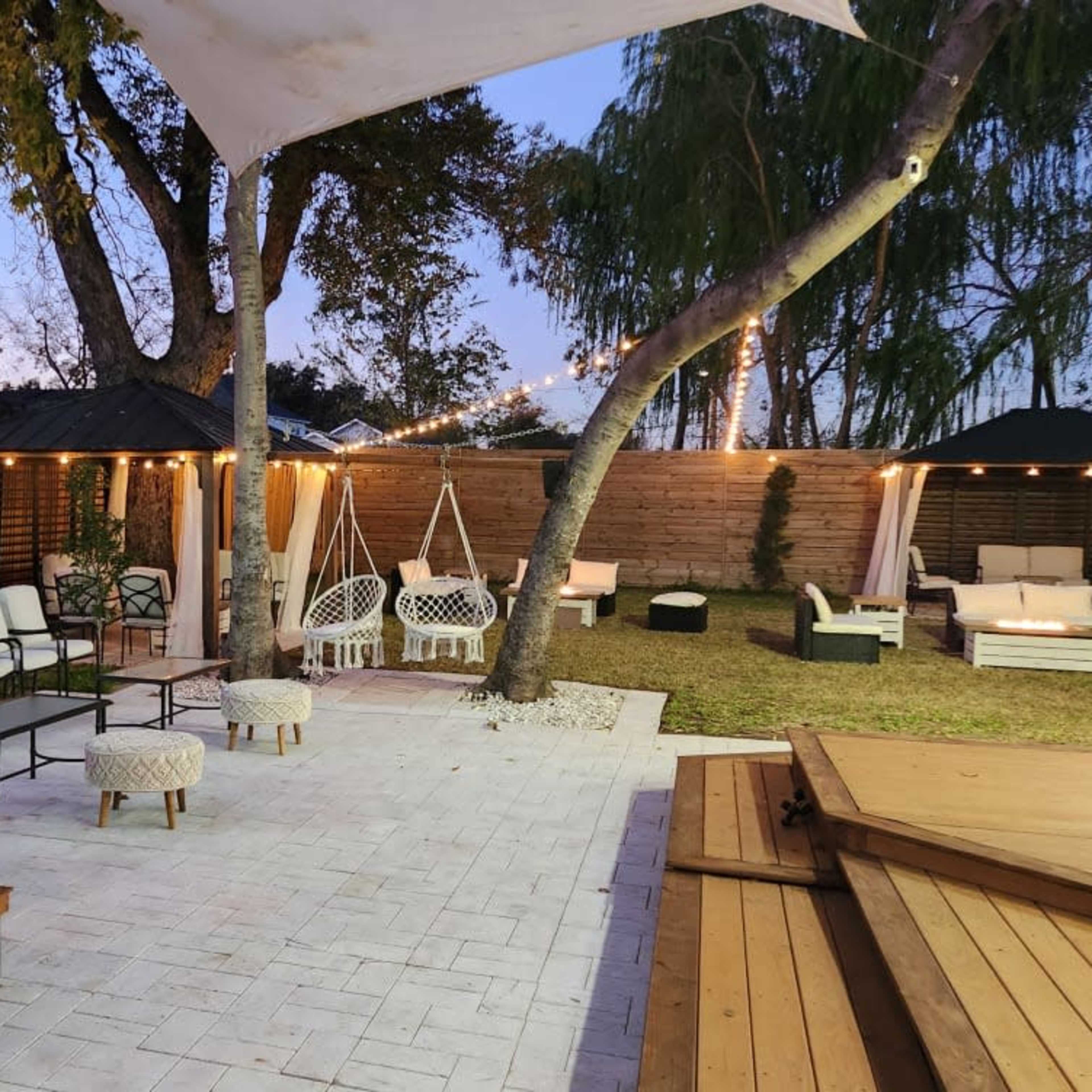 The outdoor lounge area features seating arrangements, decorative lights, and a grassy space surrounded by trees and wooden fencing.