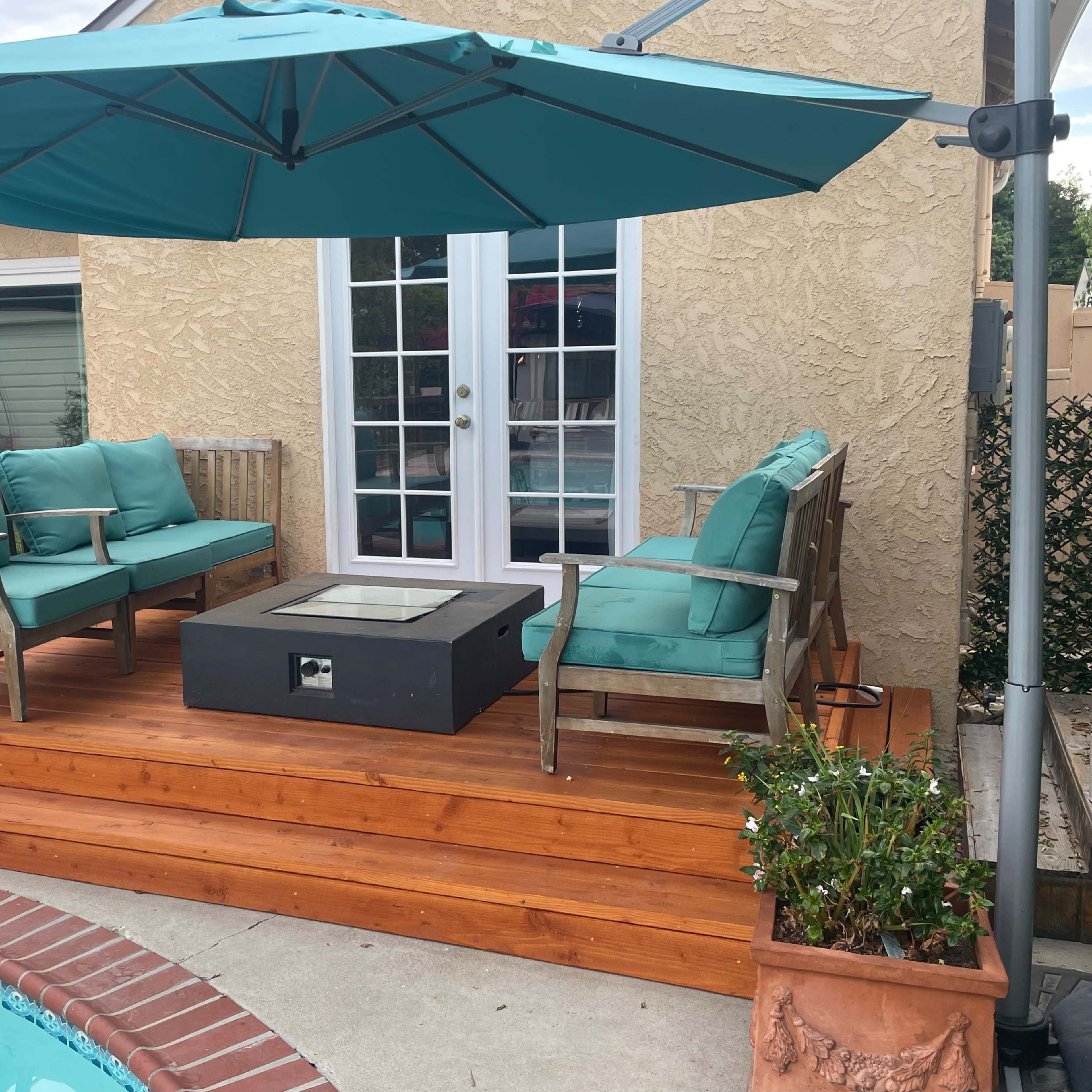 A wooden deck with a black fire table is surrounded by teal outdoor furniture and an umbrella near a swimming pool.
