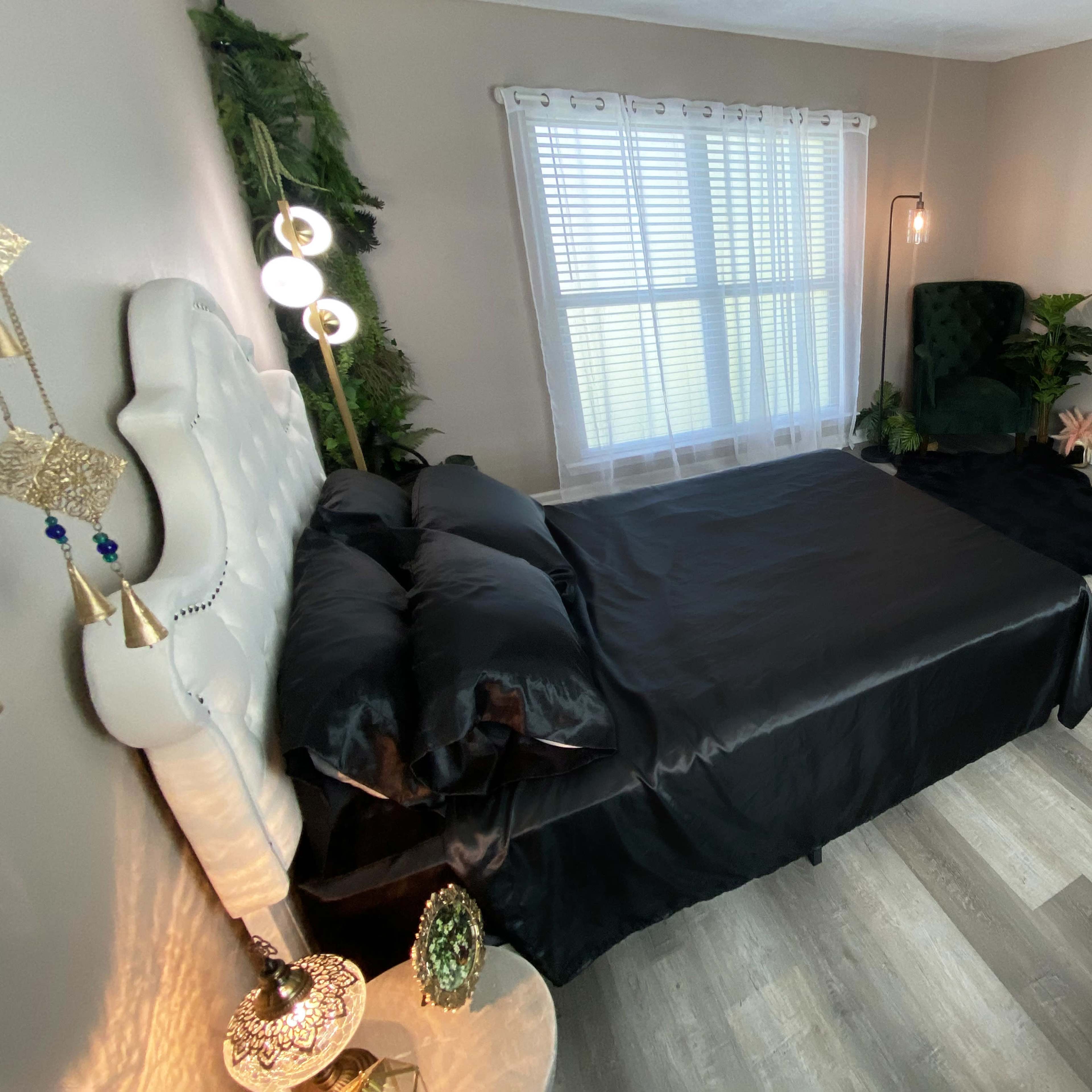 A minimalist bedroom features a black bedspread, green armchair, large mirror, and plants, with natural light filtering through sheer curtains.