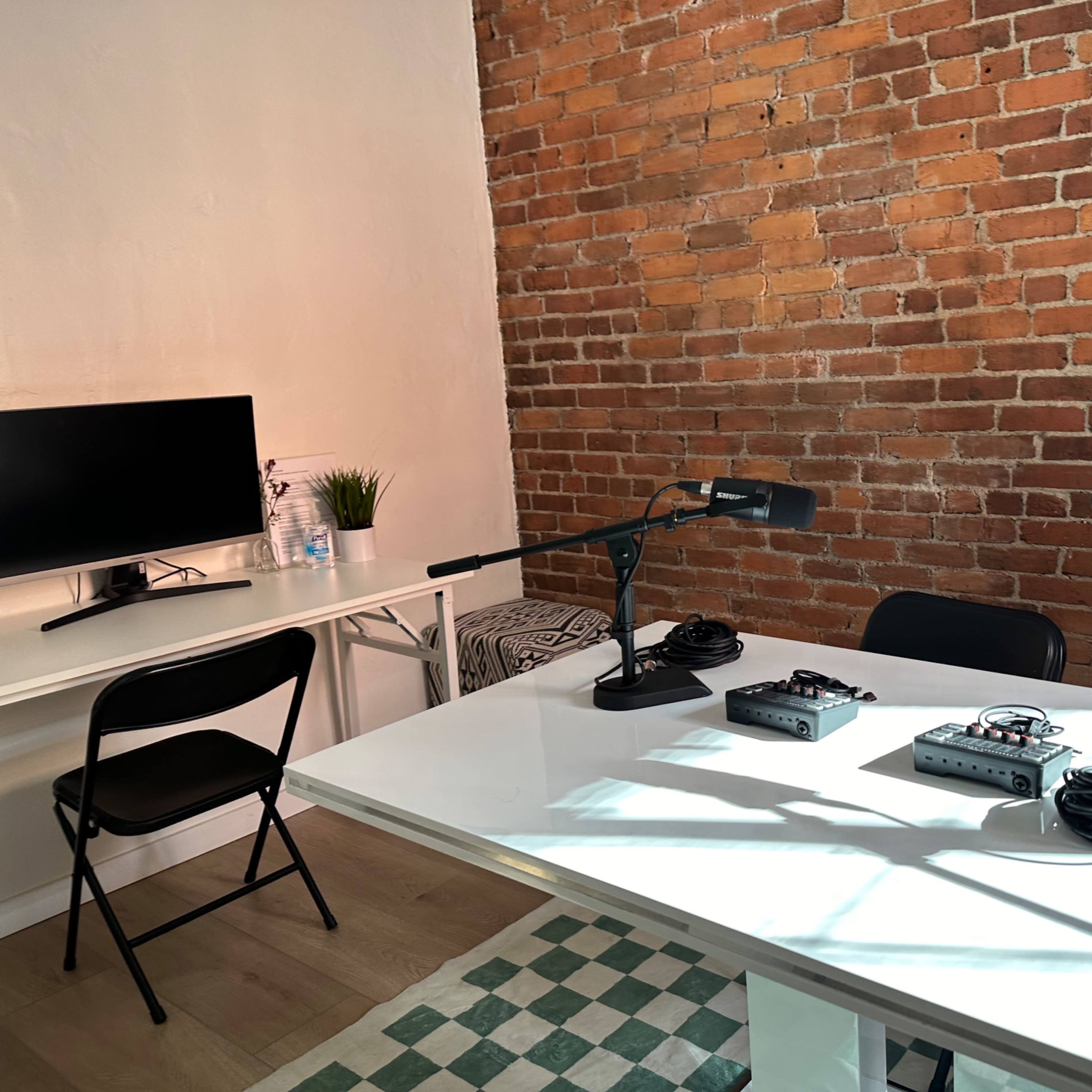 A modern recording studio with a white table, black chairs, microphones, and a brick wall.