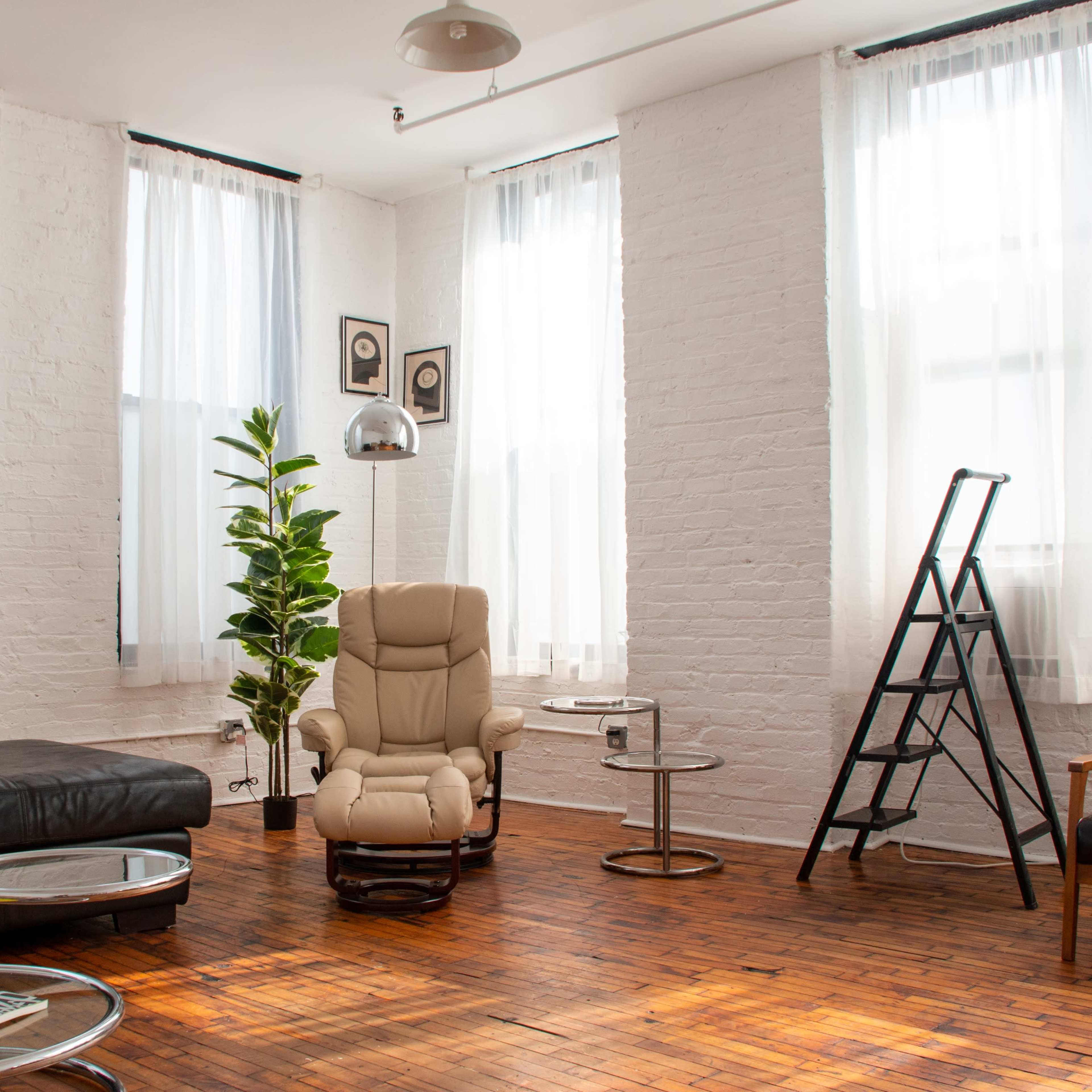 A bright room with large windows, featuring a leather sofa, a recliner chair, and a small round table on a wooden floor.