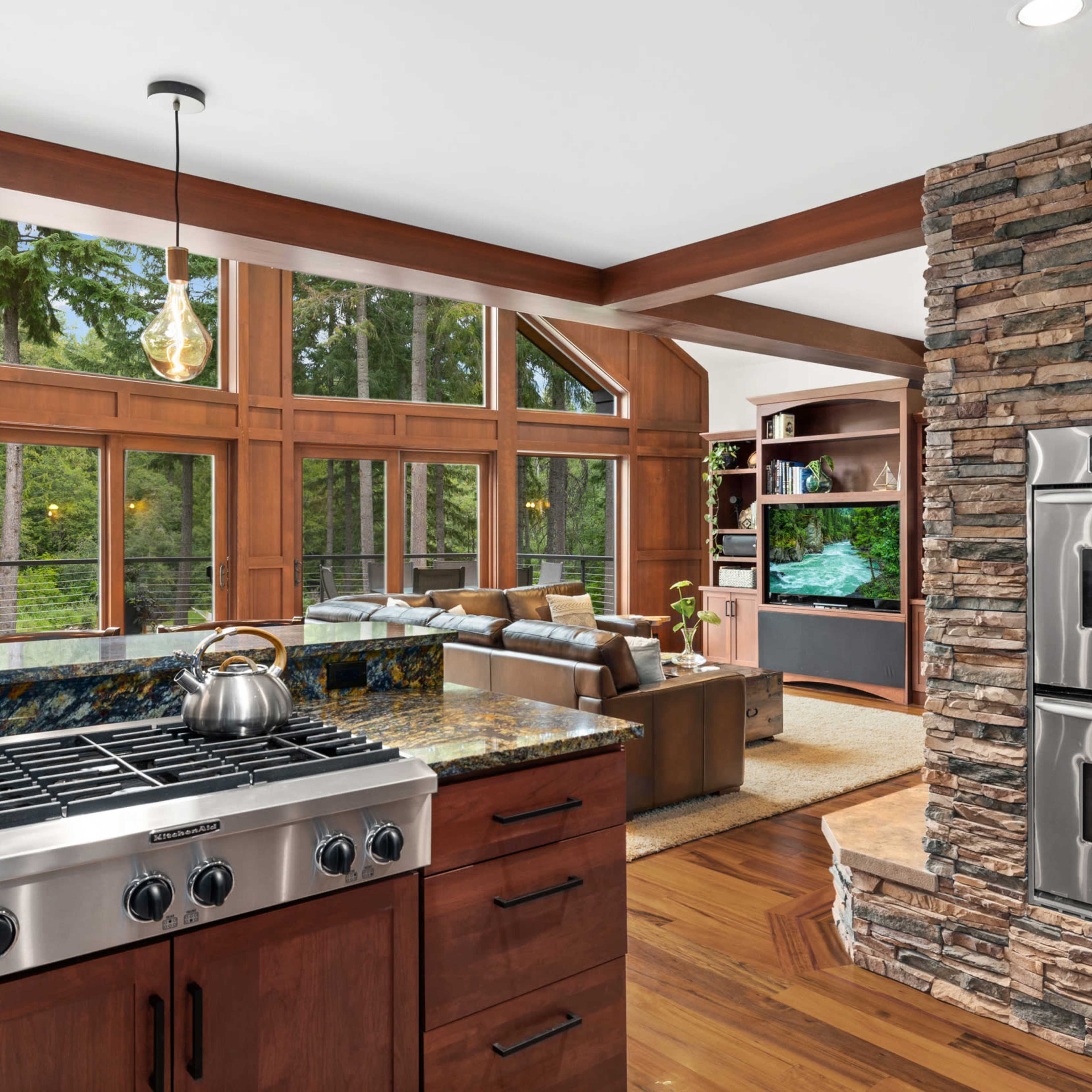 A modern kitchen features a gas stove and stainless steel double ovens, adjacent to a living area with large windows overlooking a forested landscape.