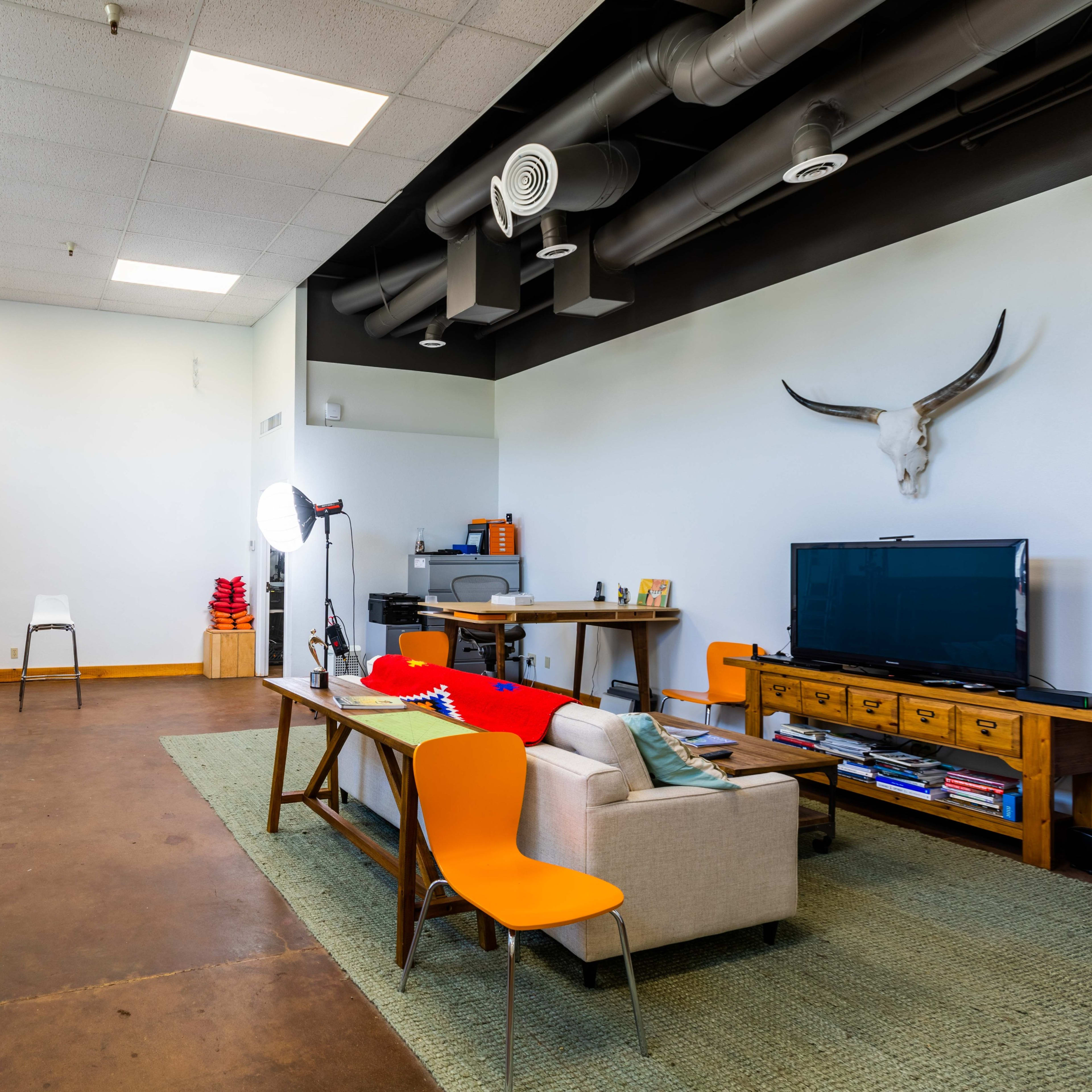 A spacious studio room furnished with a sofa, a table, a television, and a plant, illuminated by natural light from large windows.