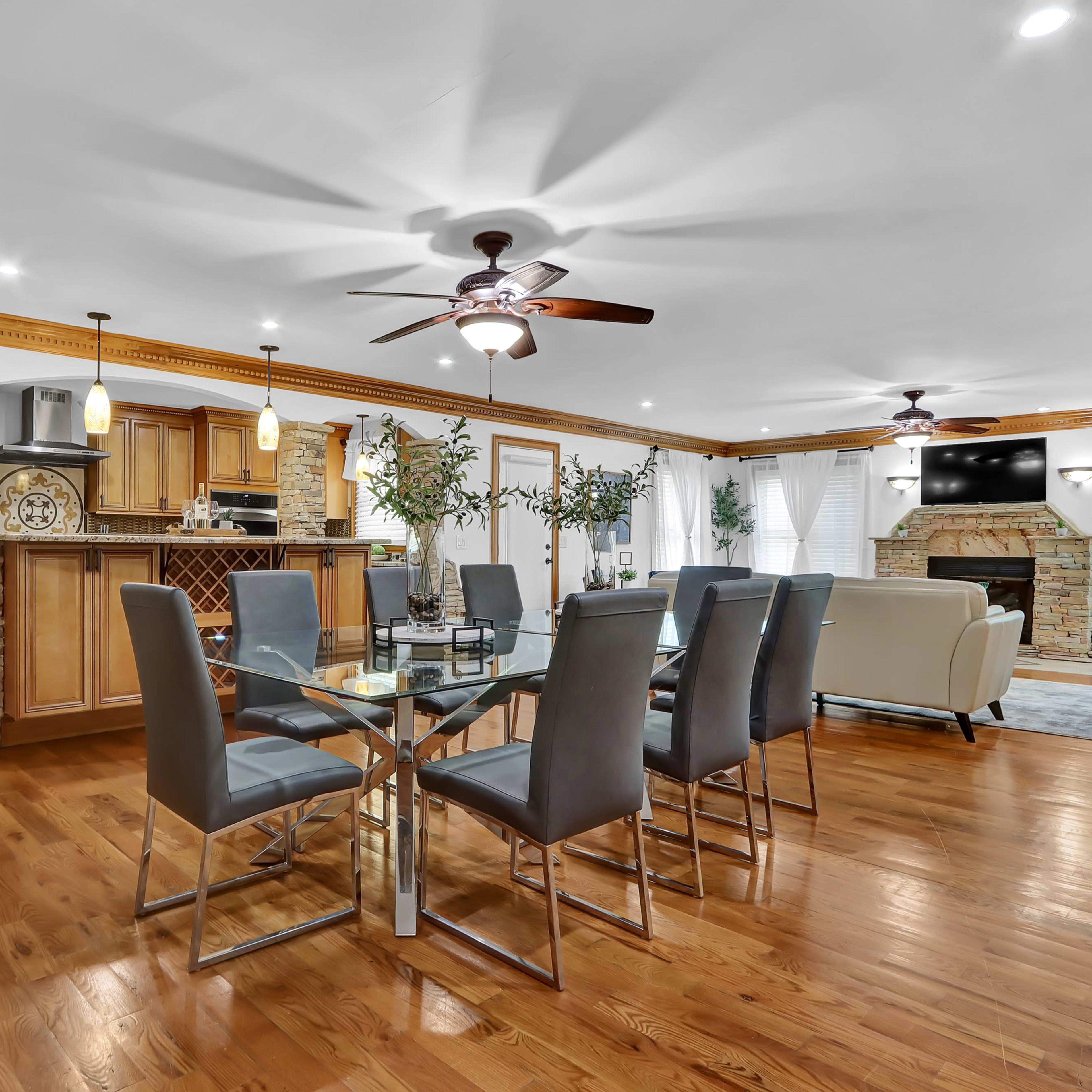 A modern, open-concept dining and living area featuring a wooden dining table with gray chairs, a stone fireplace, and a kitchen bar in the background.