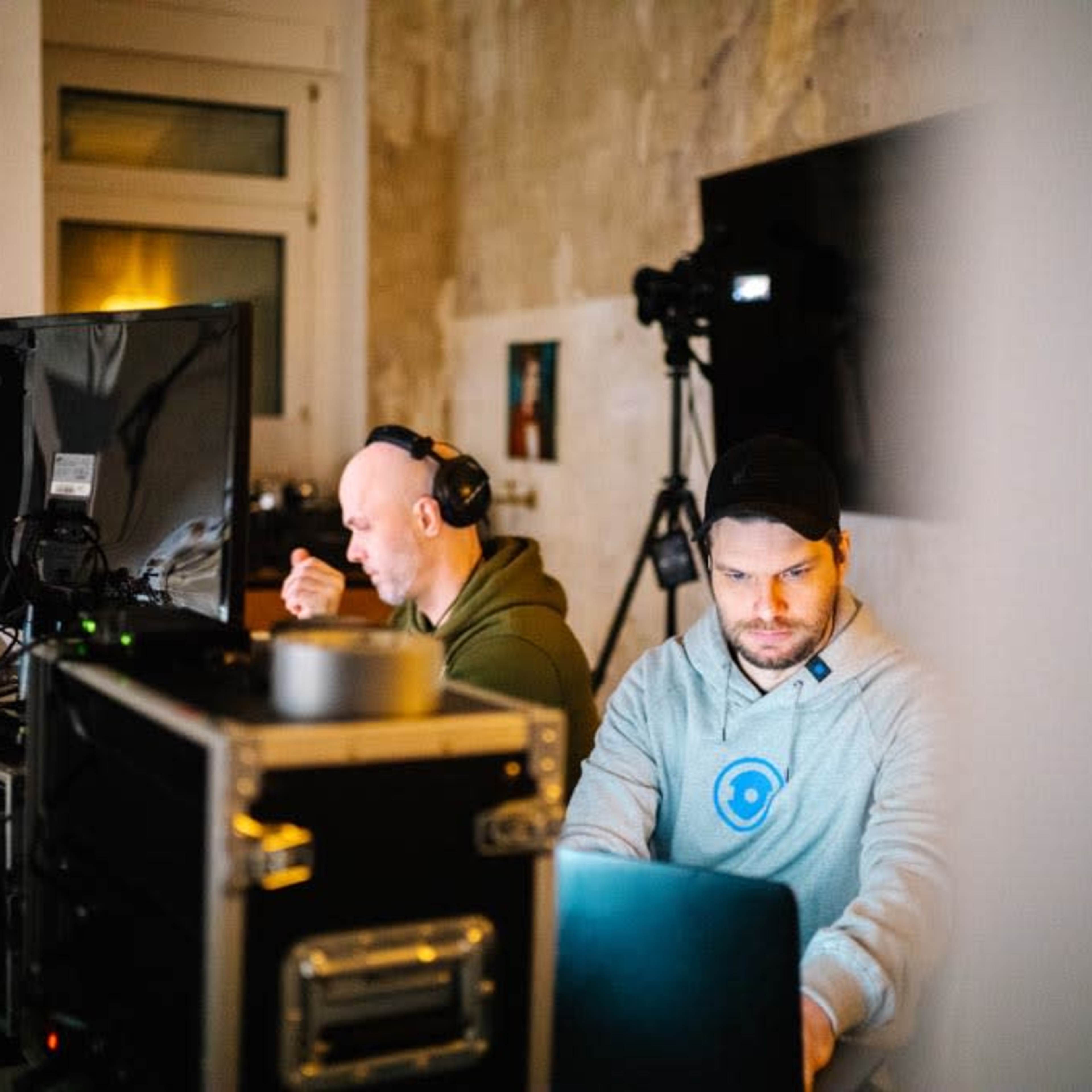 Two men work at computers in a room equipped with video production equipment and a camera.
