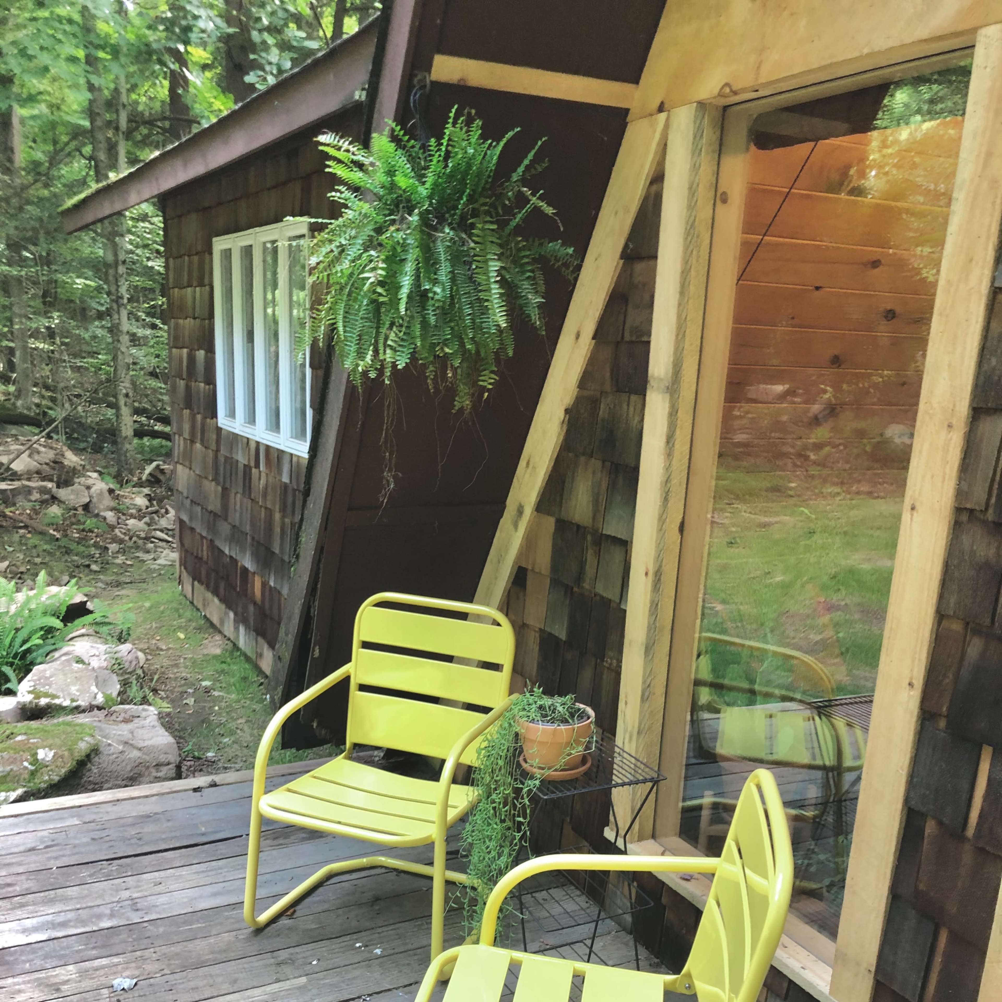 Two yellow chairs are positioned on a wooden deck beside a house with a sloped roof, framed by a hanging fern and a potted plant.