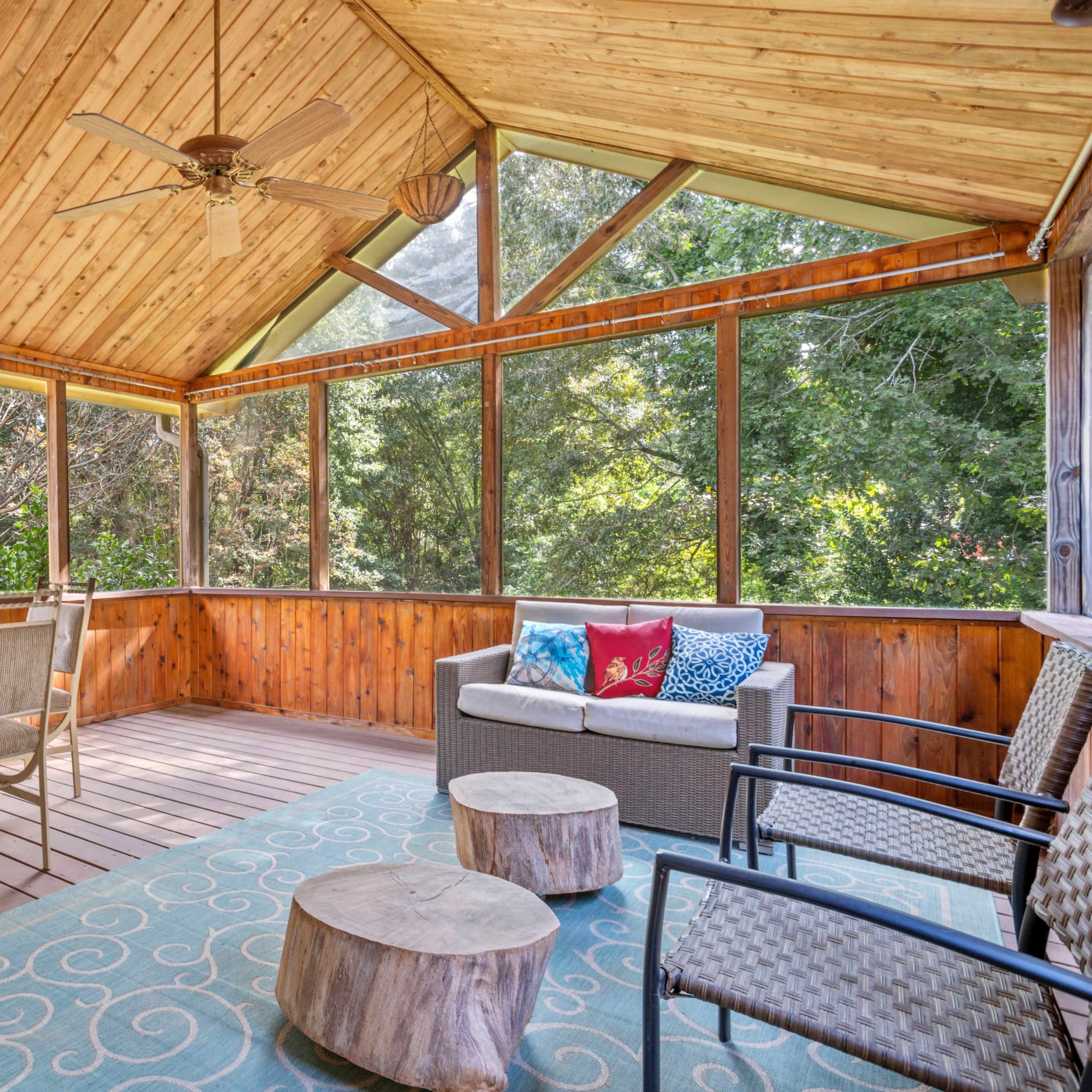 A screened-in porch with wooden walls and ceiling, featuring a dining table, seating area, and a patterned rug.