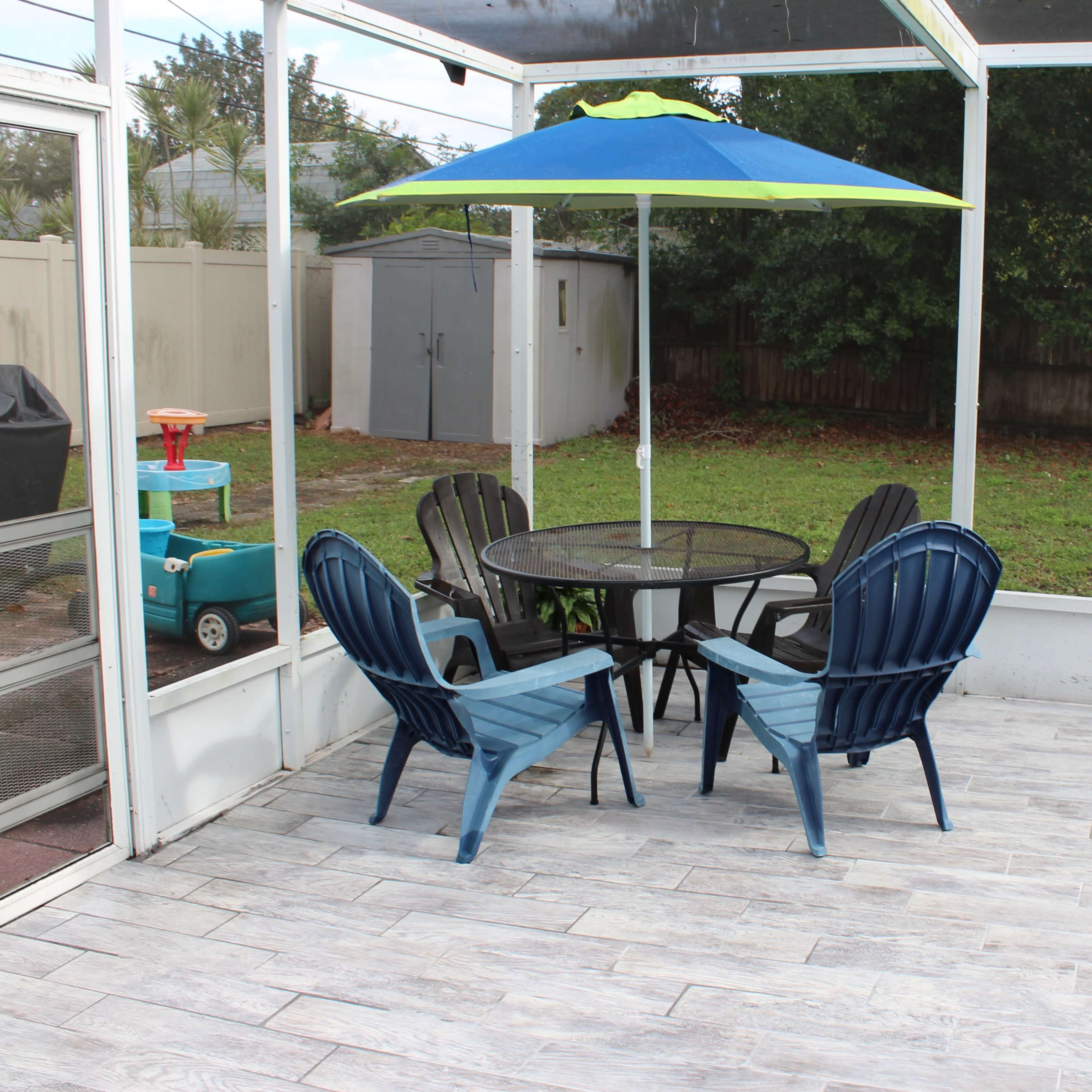A patio area with a round table surrounded by four chairs and a colorful umbrella, alongside a swimming pool and a grassy yard.