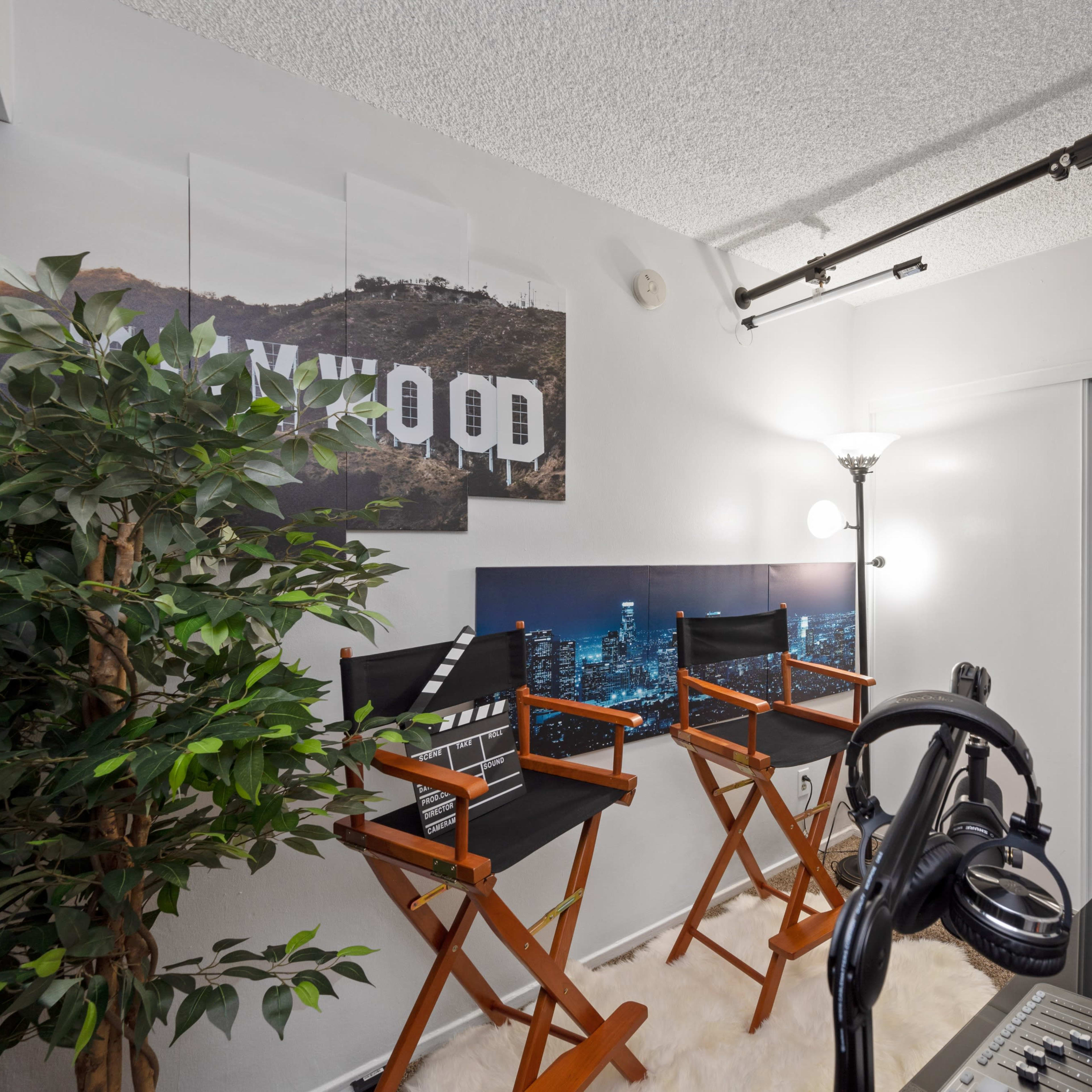 A small studio space featuring two director's chairs, a potted plant, and a microphone setup against a wall adorned with "HOLLYWOOD" signage and city skyline artwork.