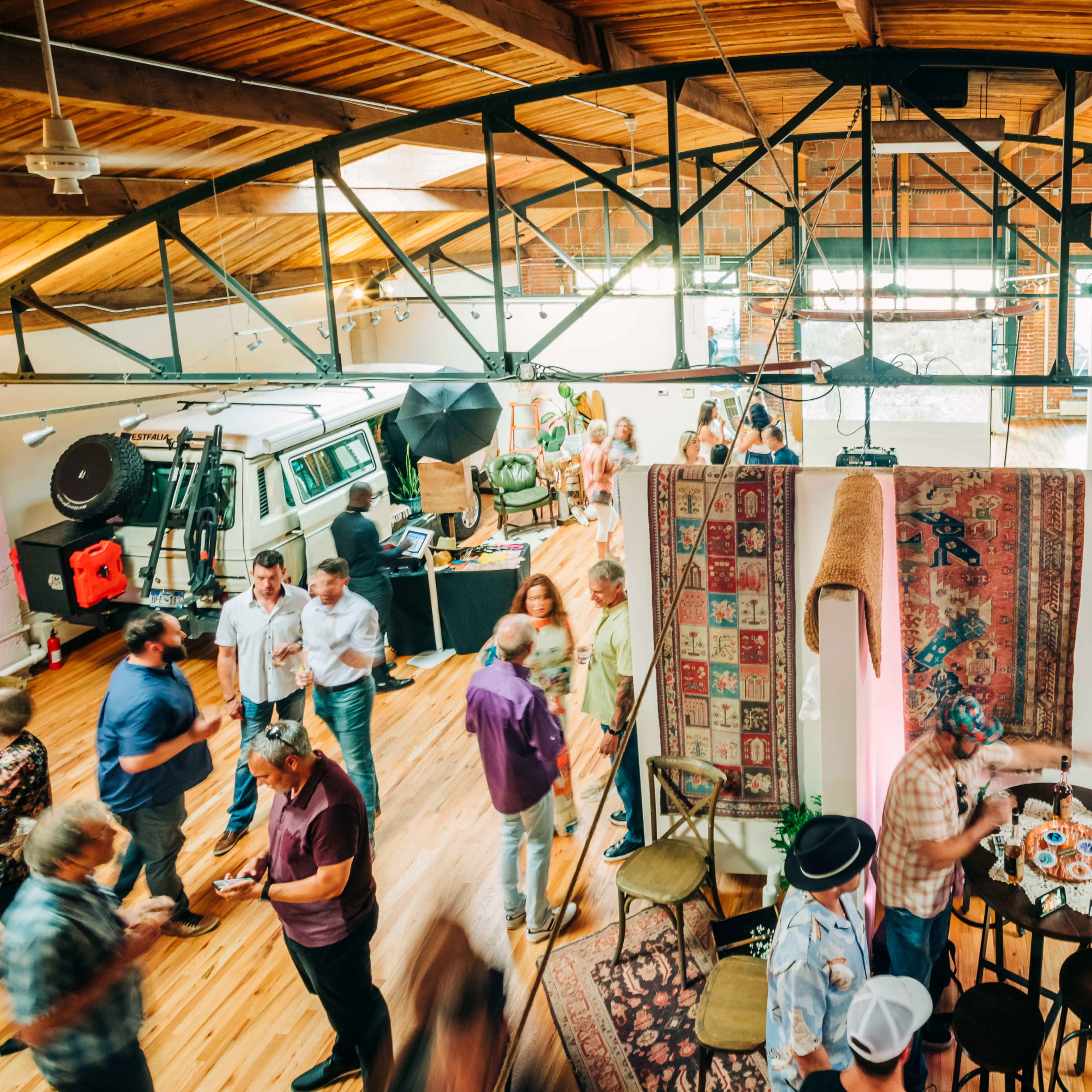 A lively gathering takes place in a spacious, brightly lit venue with attendees mingling around tables and displays.
