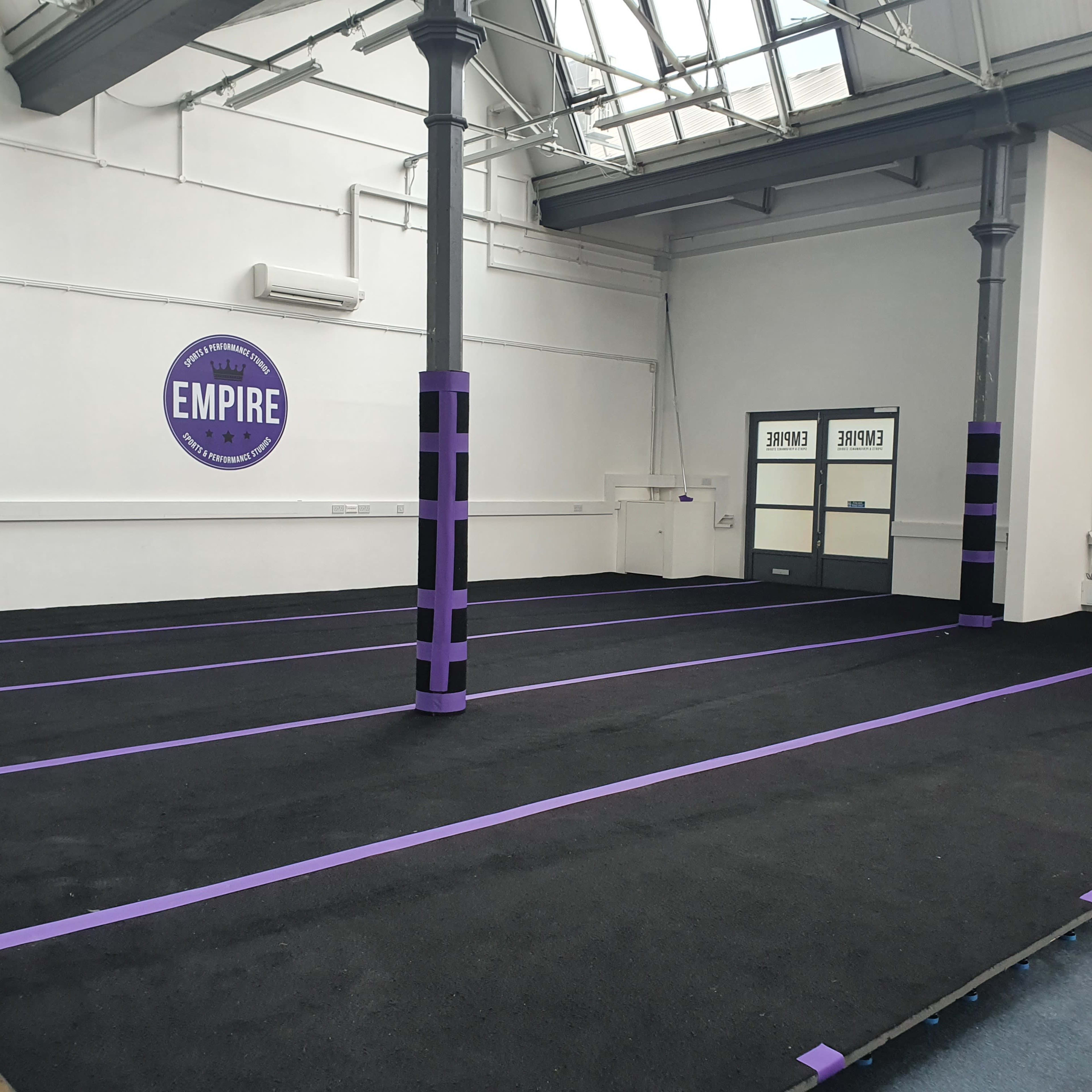 A spacious indoor area with a black floor, marked by purple tape, and features tall pillars, one of which has an "EMPIRE" logo on the wall.