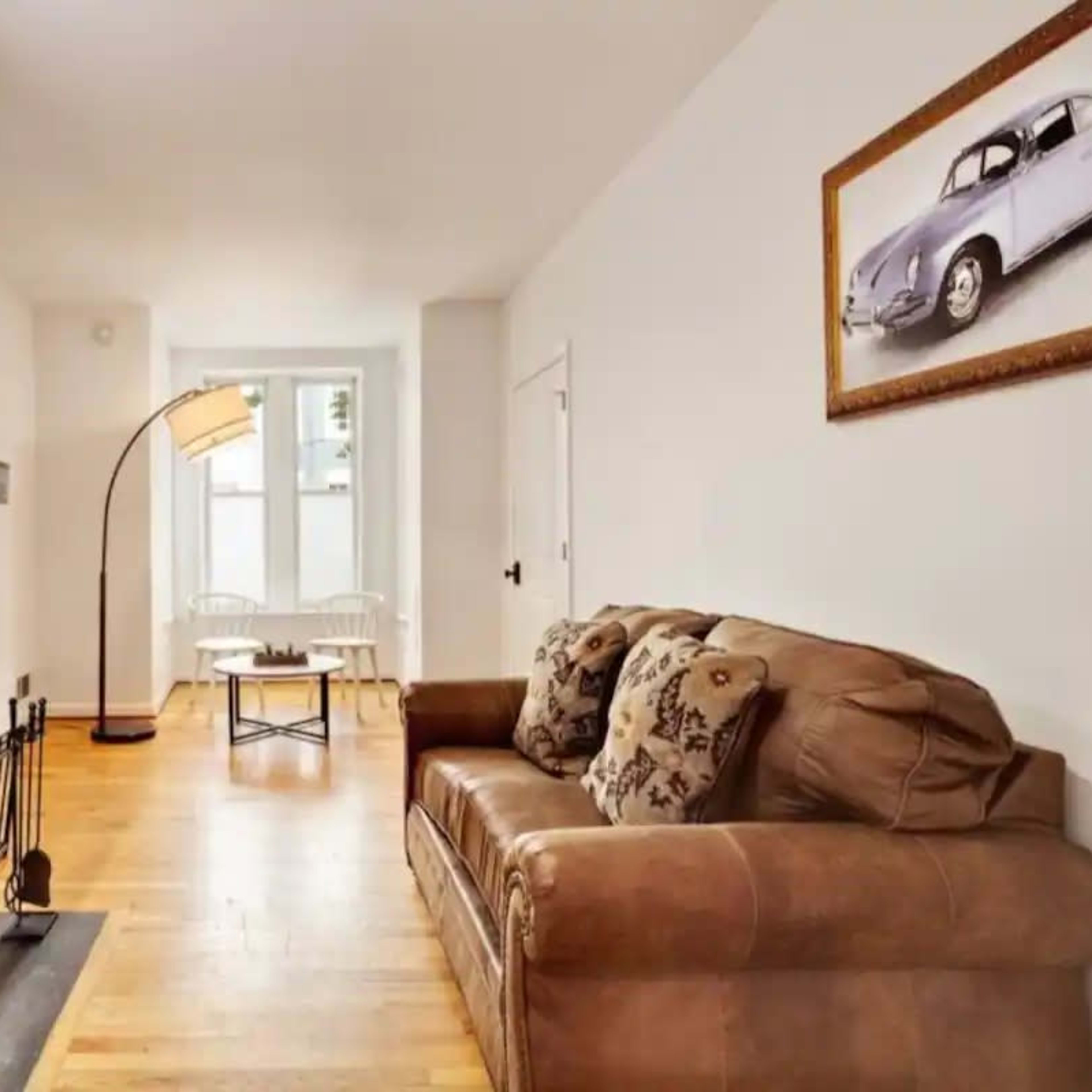 A living room with a brown leather sofa, a fireplace, a floor lamp, and a framed picture of a vintage car on the wall.