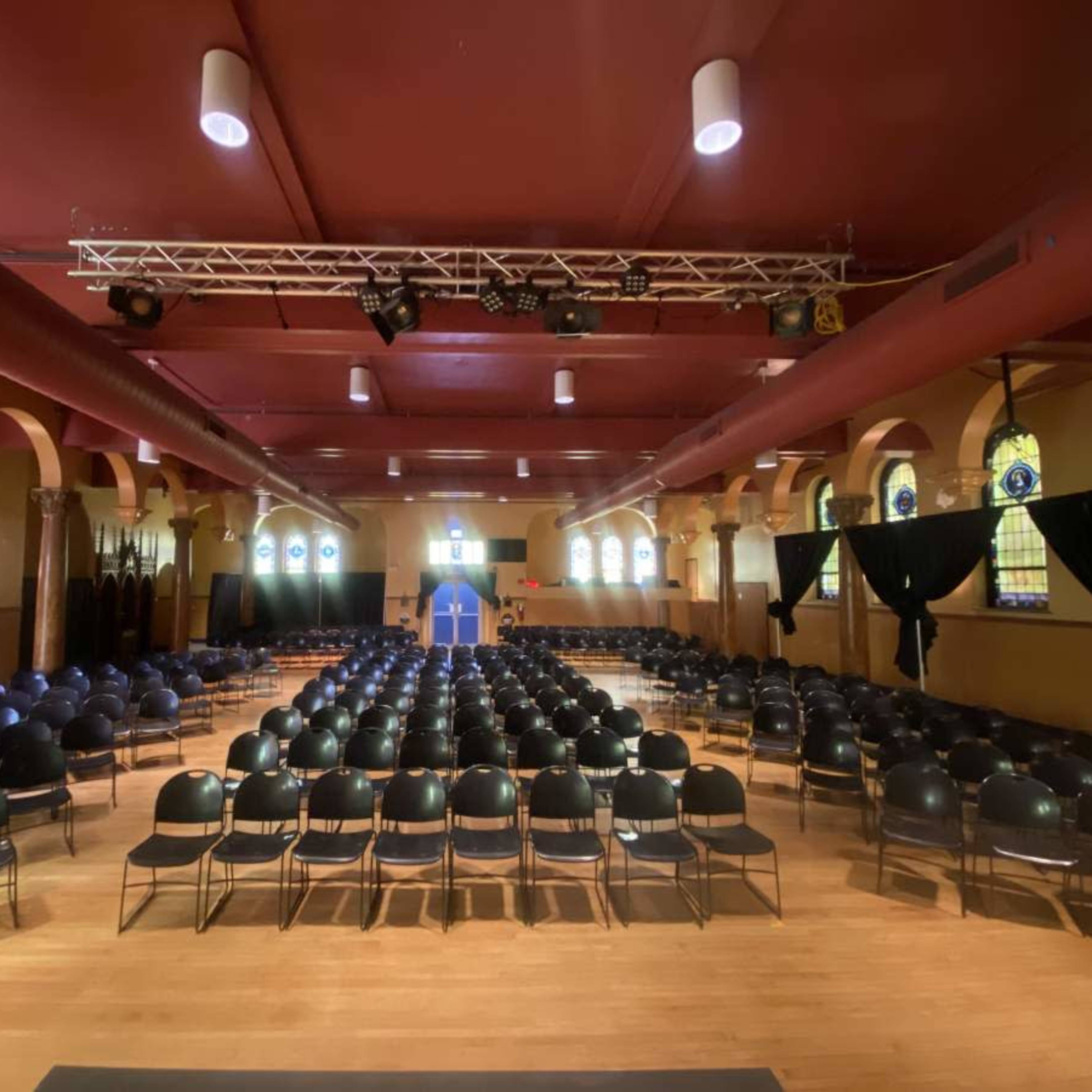 A spacious hall arranged with rows of black chairs facing a central stage, illuminated by overhead lights, with stained glass windows in the background.