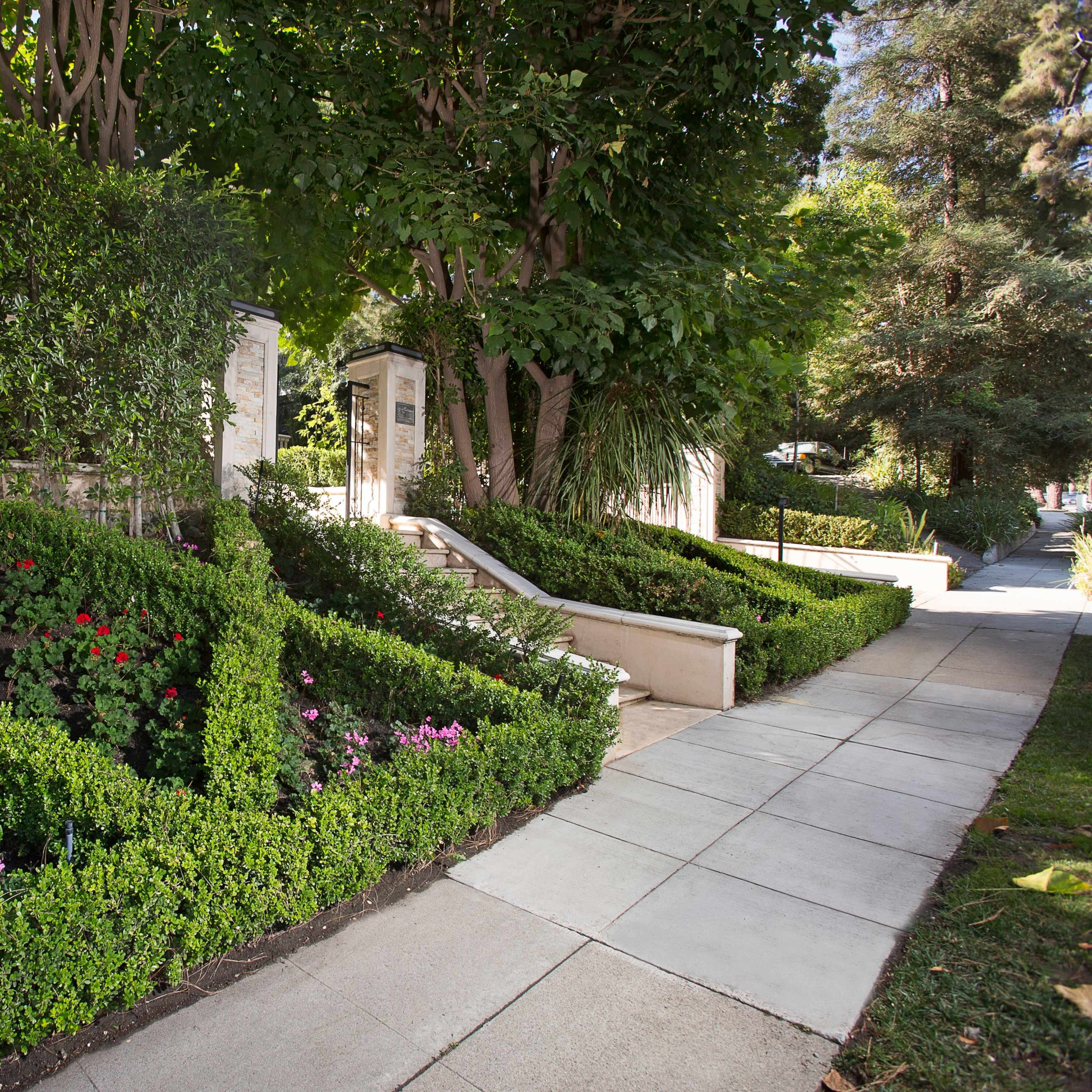 A landscaped pathway bordered by neatly trimmed hedges and flowerbeds, leading towards a set of steps and garden features.