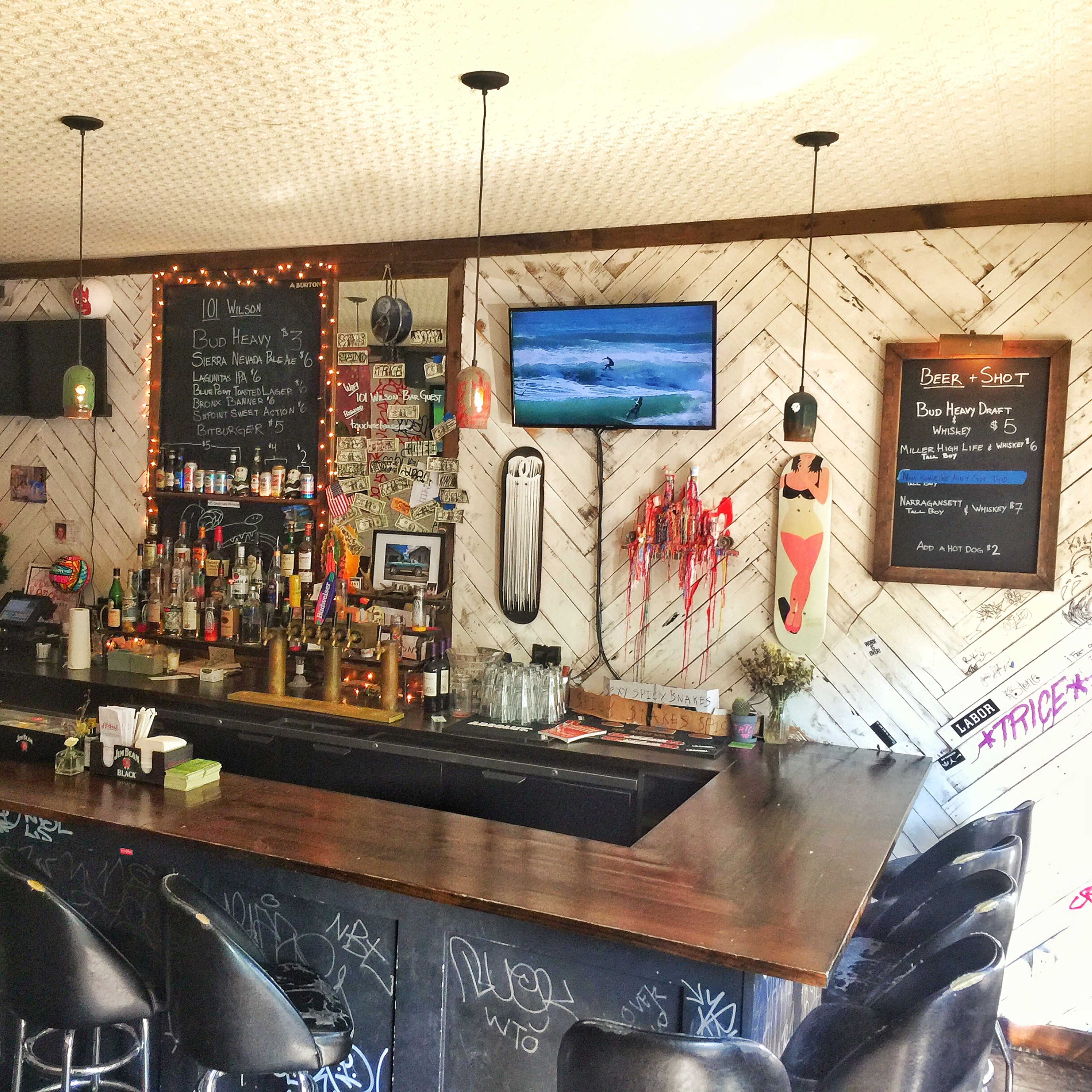 A bar area with a dark wood counter, shelves of bottles, and a TV displaying a surfing scene, surrounded by festive decorations and graffiti on the walls.