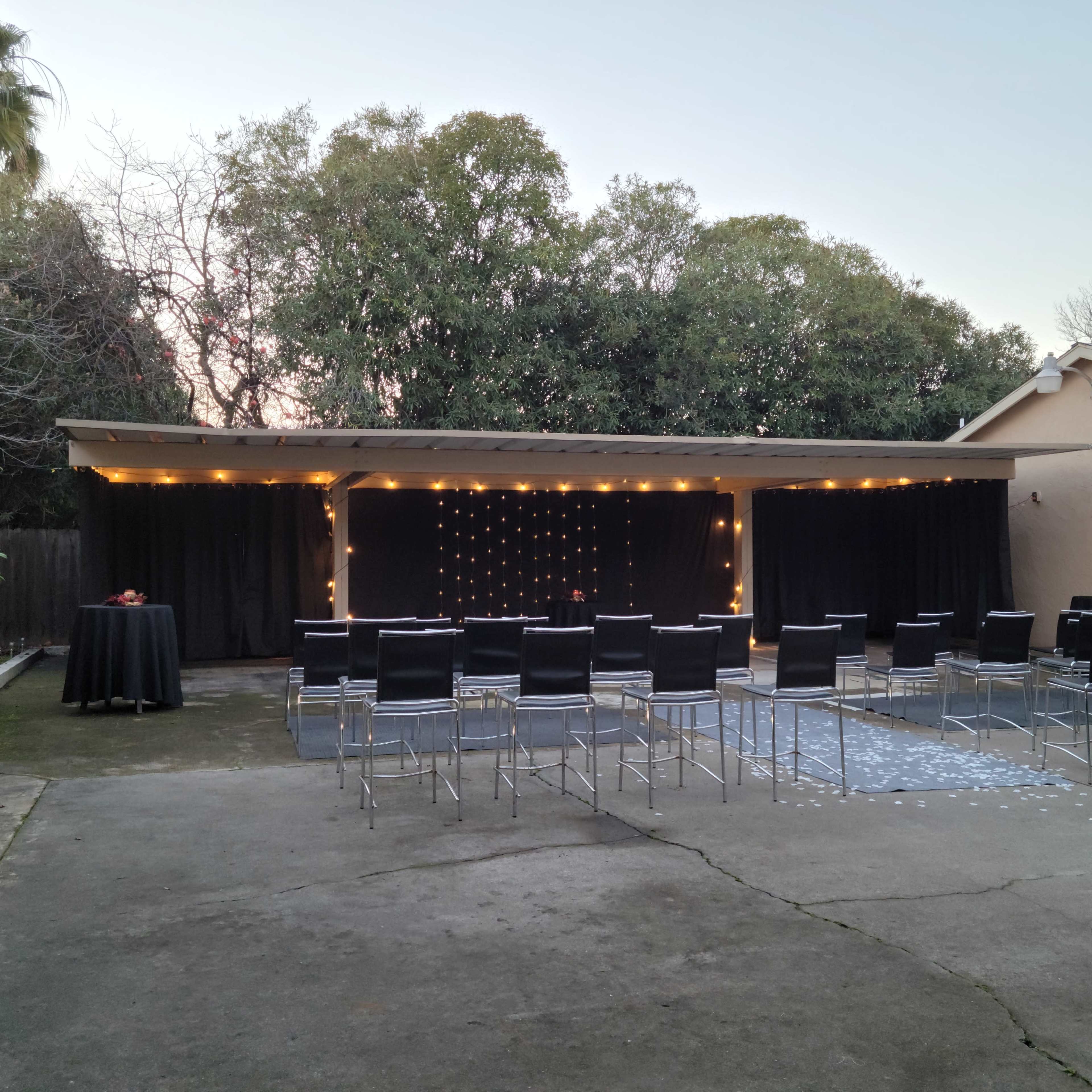 An outdoor space featuring a stage with black curtains, decorated with string lights, and arranged seating in front.