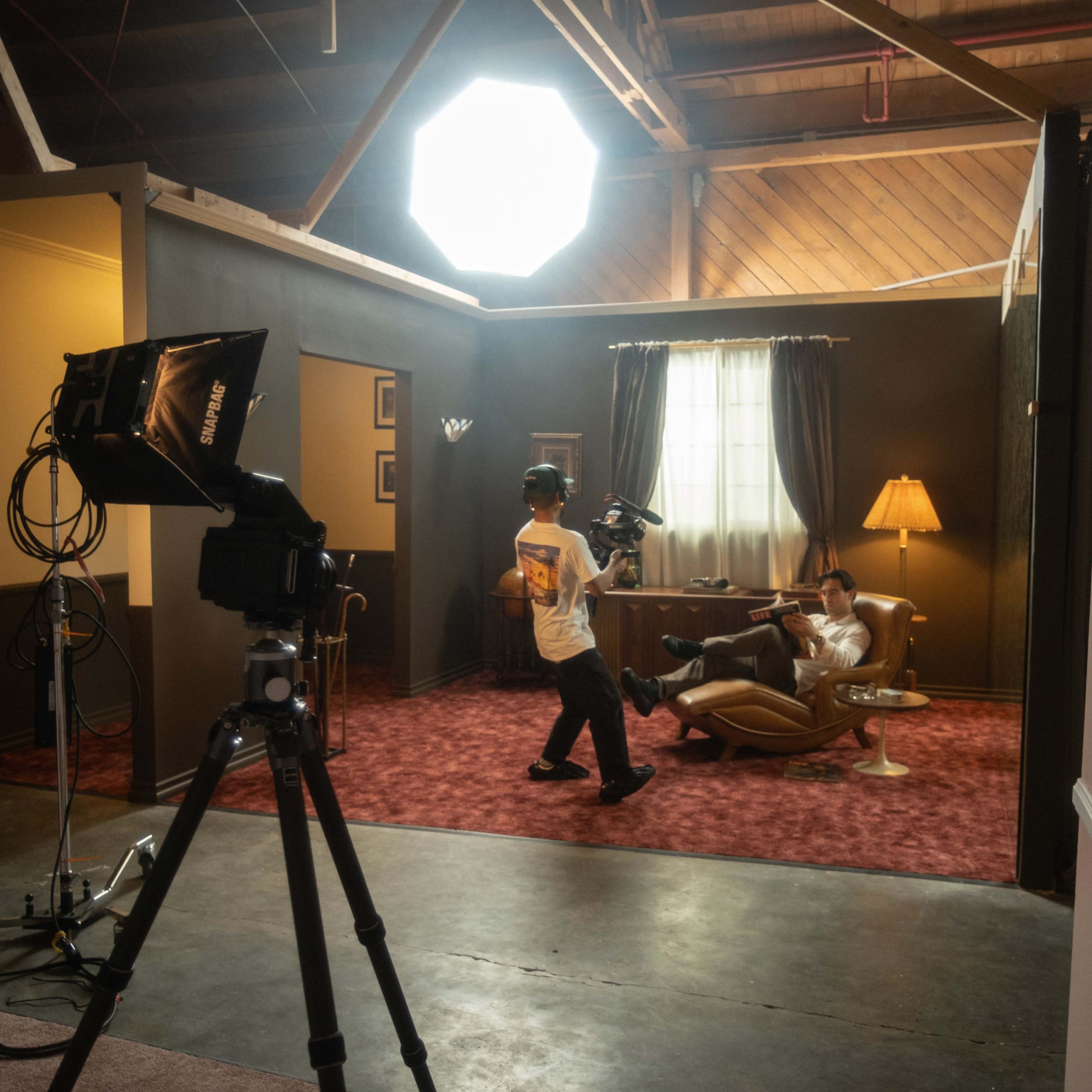 A filmmaker operates a camera while a subject relaxes in an armchair, with studio lighting illuminating a vintage-style interior.