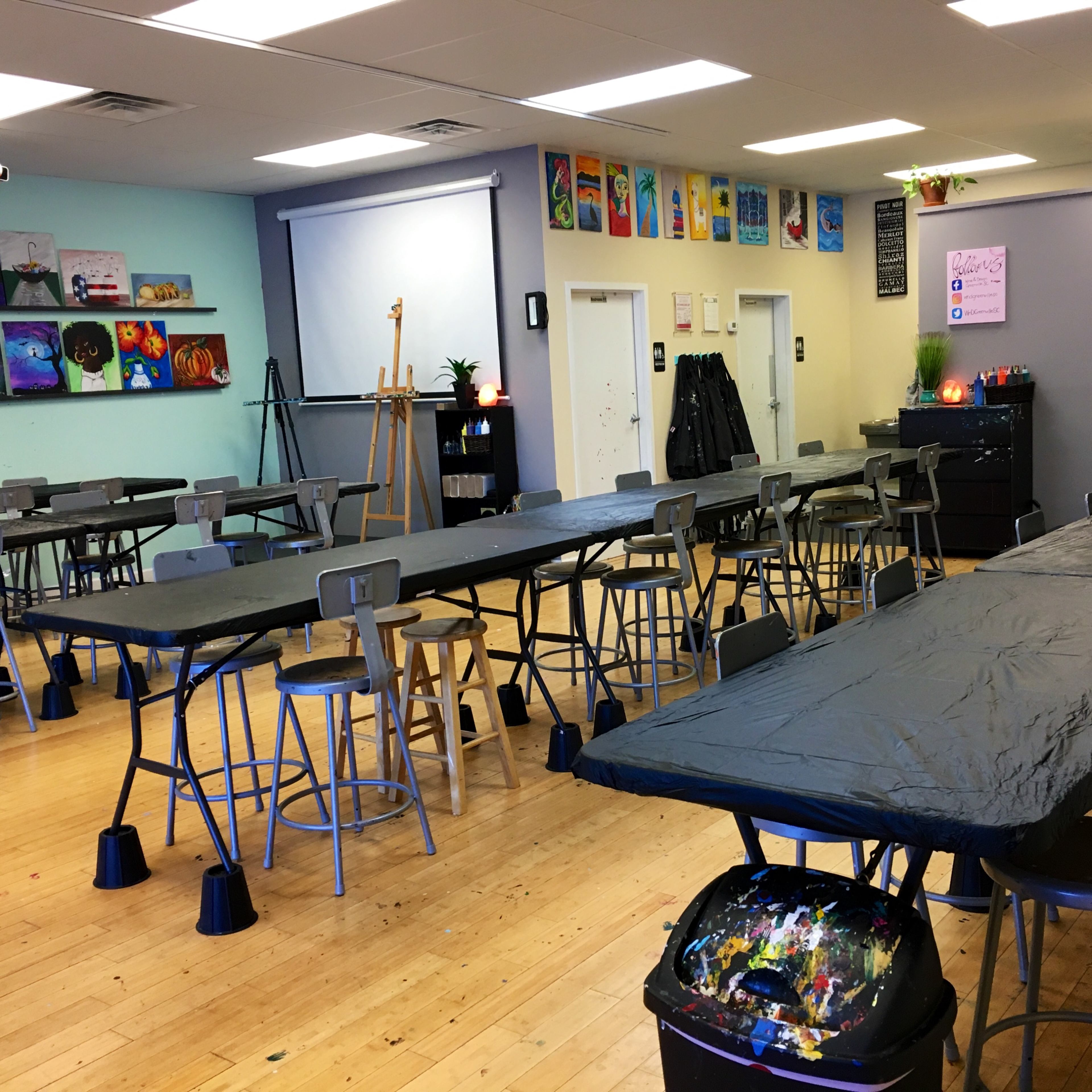 An art classroom with several long tables covered in black tablecloths, surrounded by stools and featuring easels and colorful artwork on the walls.