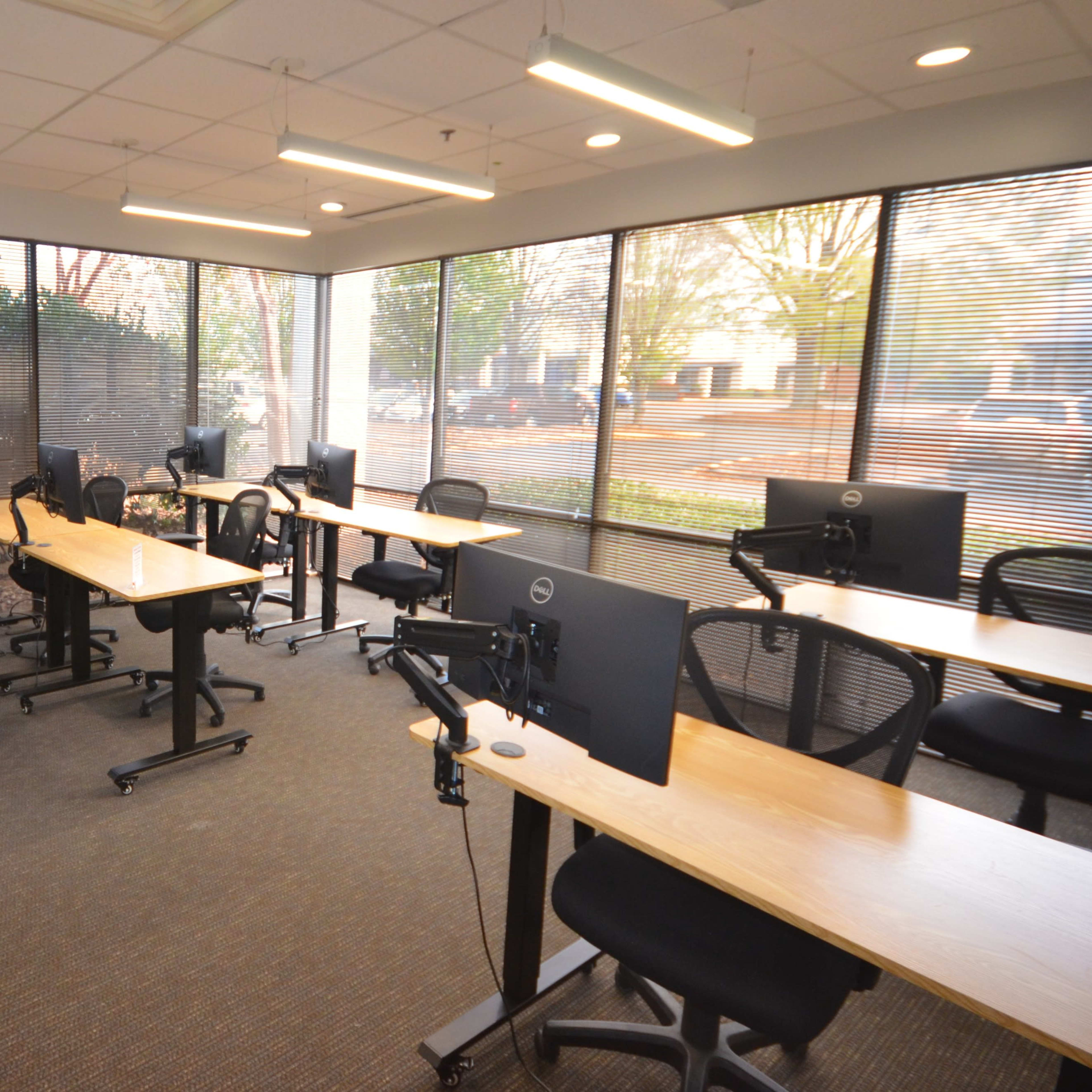 A modern classroom with several desks and computer monitors, surrounded by large windows that provide natural light.