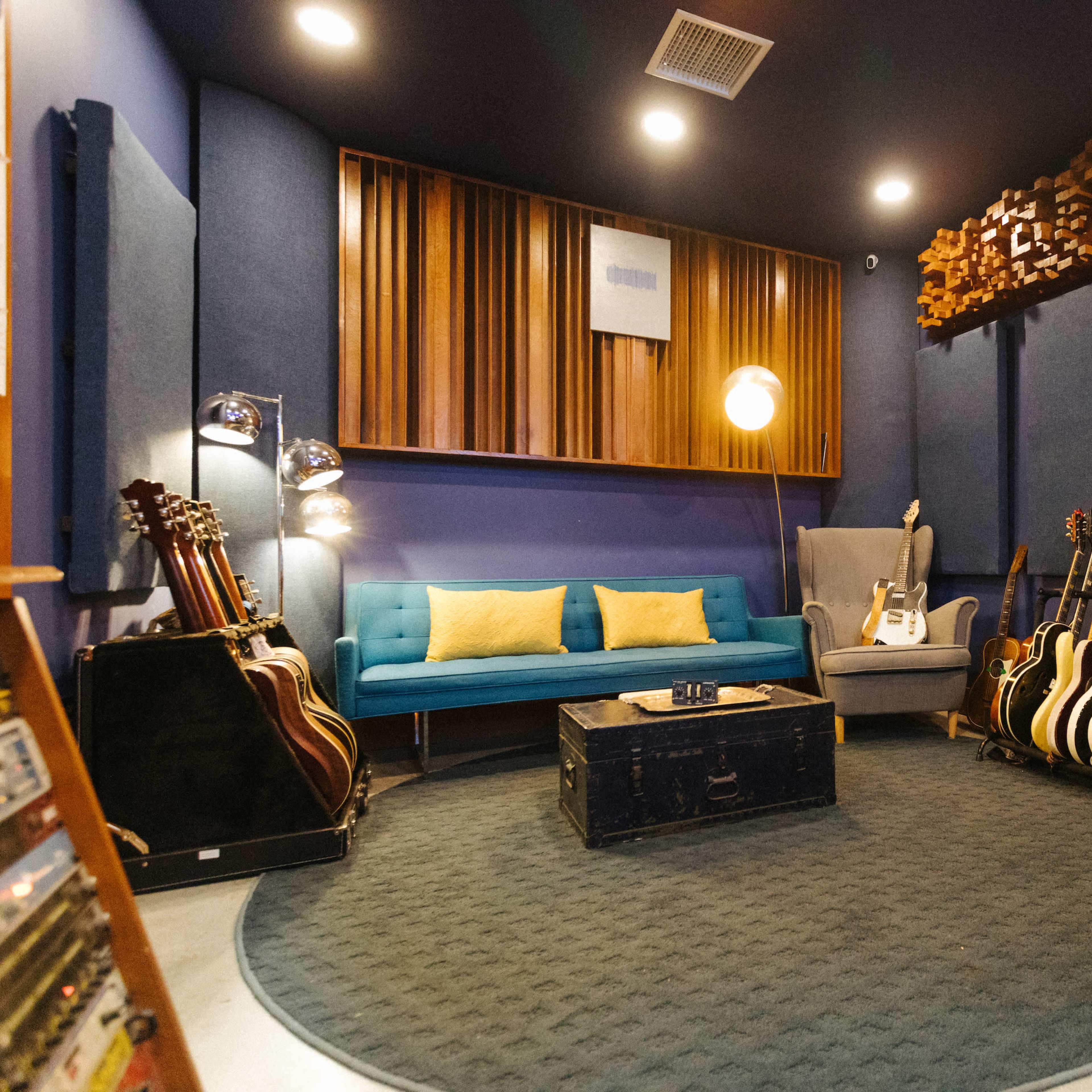 A music studio features a blue couch, a gray armchair, a round rug, a collection of guitars, and soundproofing panels on the walls.